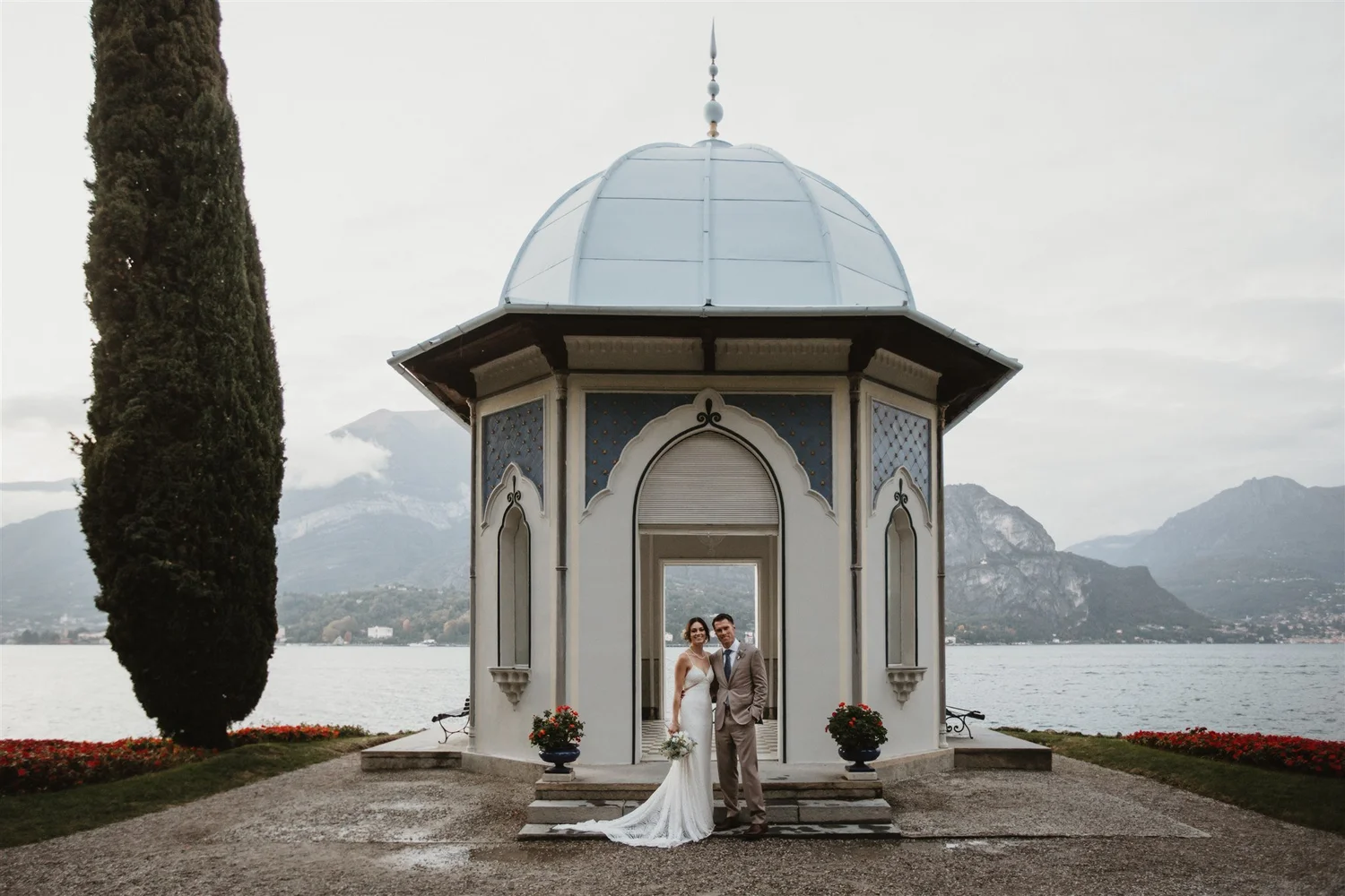 A bride and groom stand together under a decorative gazebo overlooking a lake surrounded by mountains, with cloudy skies overhead. The bride is wearing a white wedding dress, and the groom is in a light-colored suit.