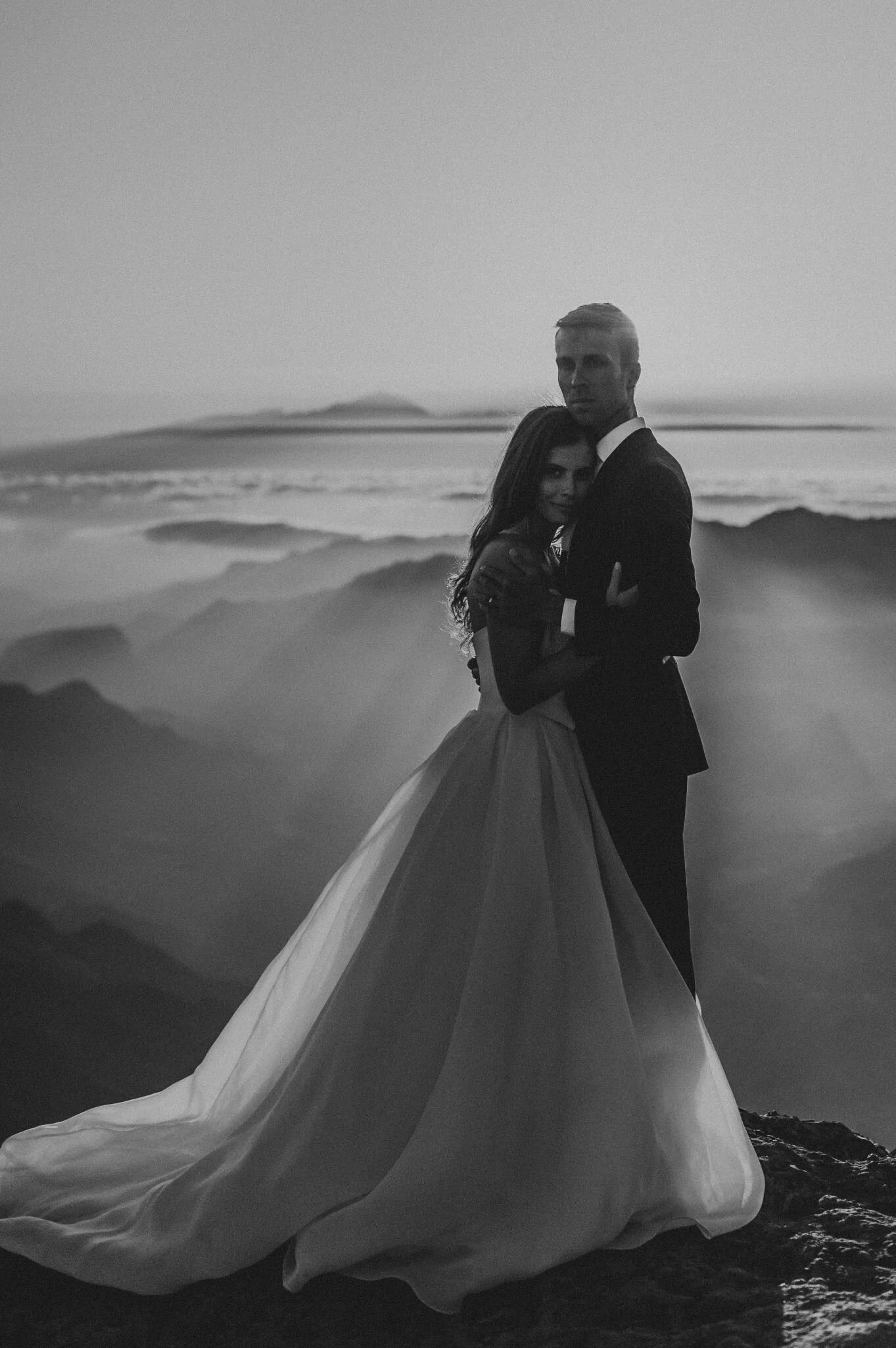 A black and white photo of a bride and groom embracing on a beach at sunset. The bride wears a flowing wedding dress, and the groom wears a suit.