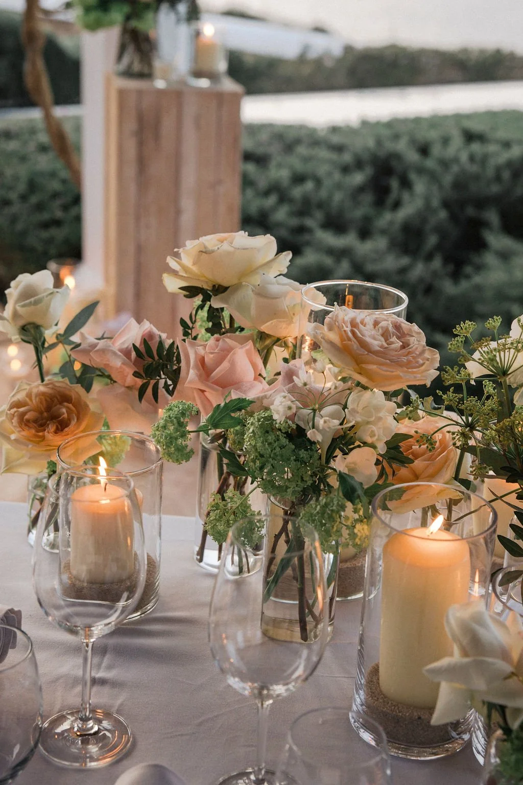 A table decorated with a floral arrangement of pink and white roses, green foliage, and candles in glass holders, set outdoors with a view of a body of water and greenery in the background.