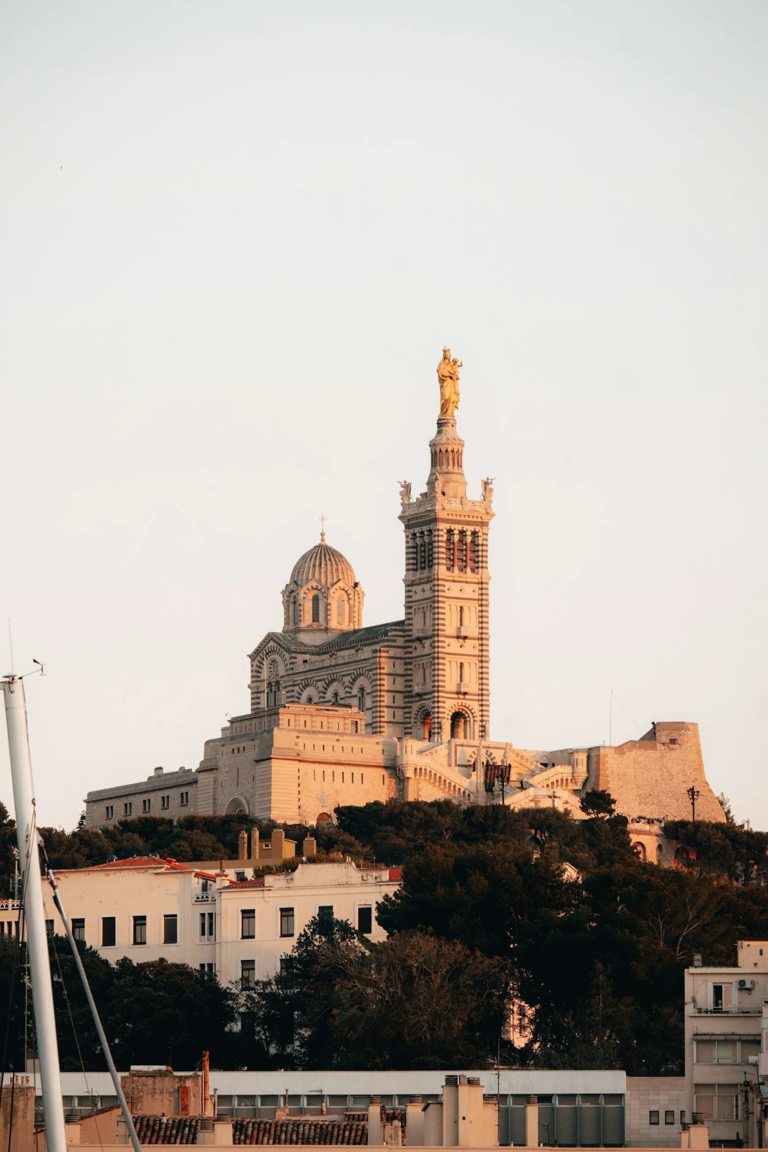 A tall church or cathedral with a large dome and a steeple topped with a golden statue, situated on a hill and illuminated by warm sunlight with a clear sky background.