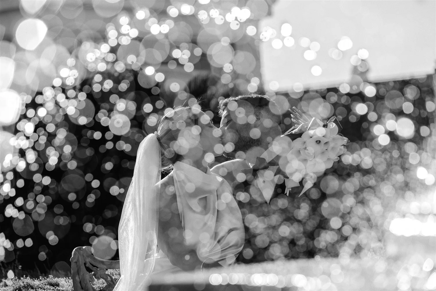 A couple kissing, surrounded by bokeh light effects, with a bouquet of flowers on the side, in a black and white photograph.