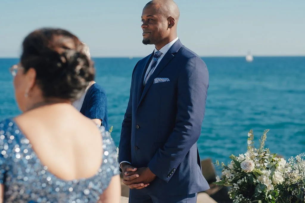 A man in a navy suit and a woman in a sparkly dress at a seaside wedding ceremony with flowers and the ocean in the background.