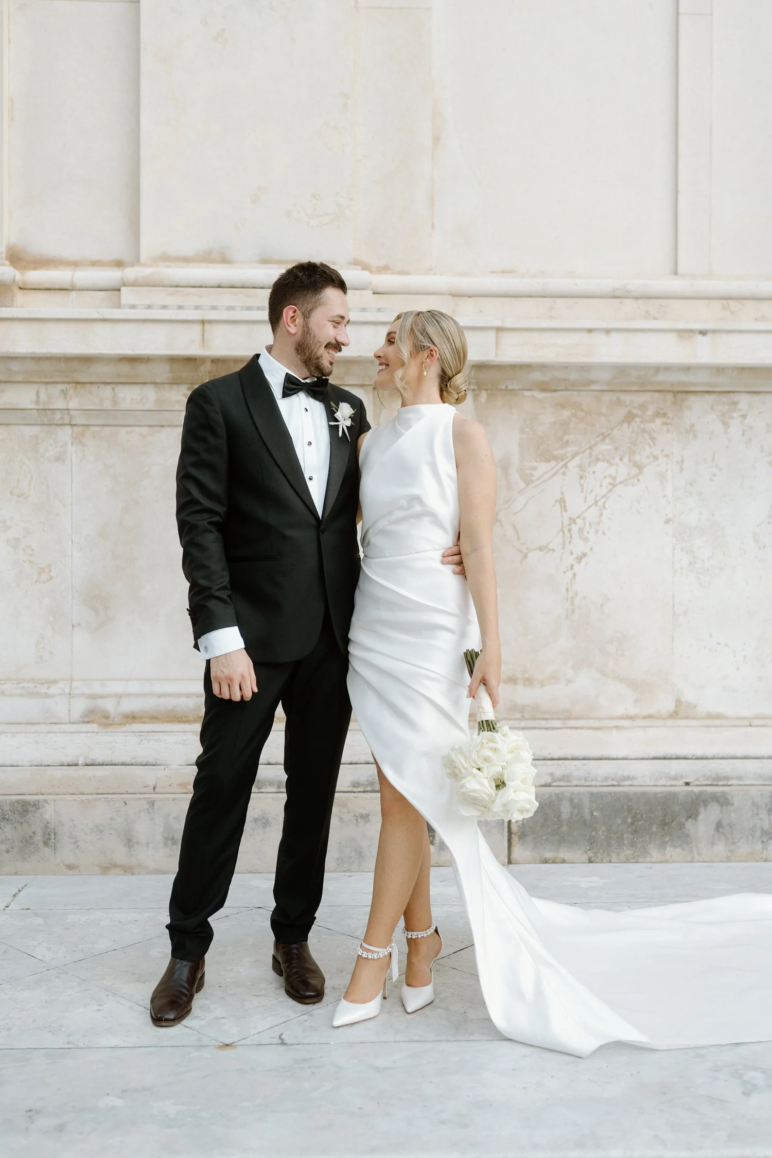A bride and groom stand close together, smiling and looking at each other, in front of a large stone building with steps. The groom wears a black tuxedo with a bow tie, and the bride wears a sleeveless white wedding dress with a train, holding a bouq