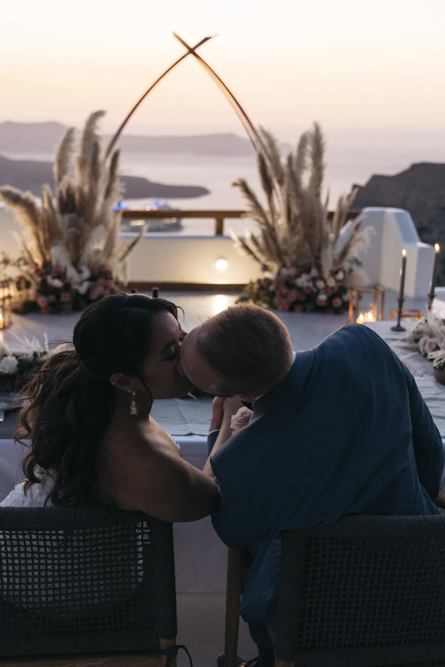 A couple kissing during sunset at an outdoor wedding ceremony with decorative floral arrangements and candles.