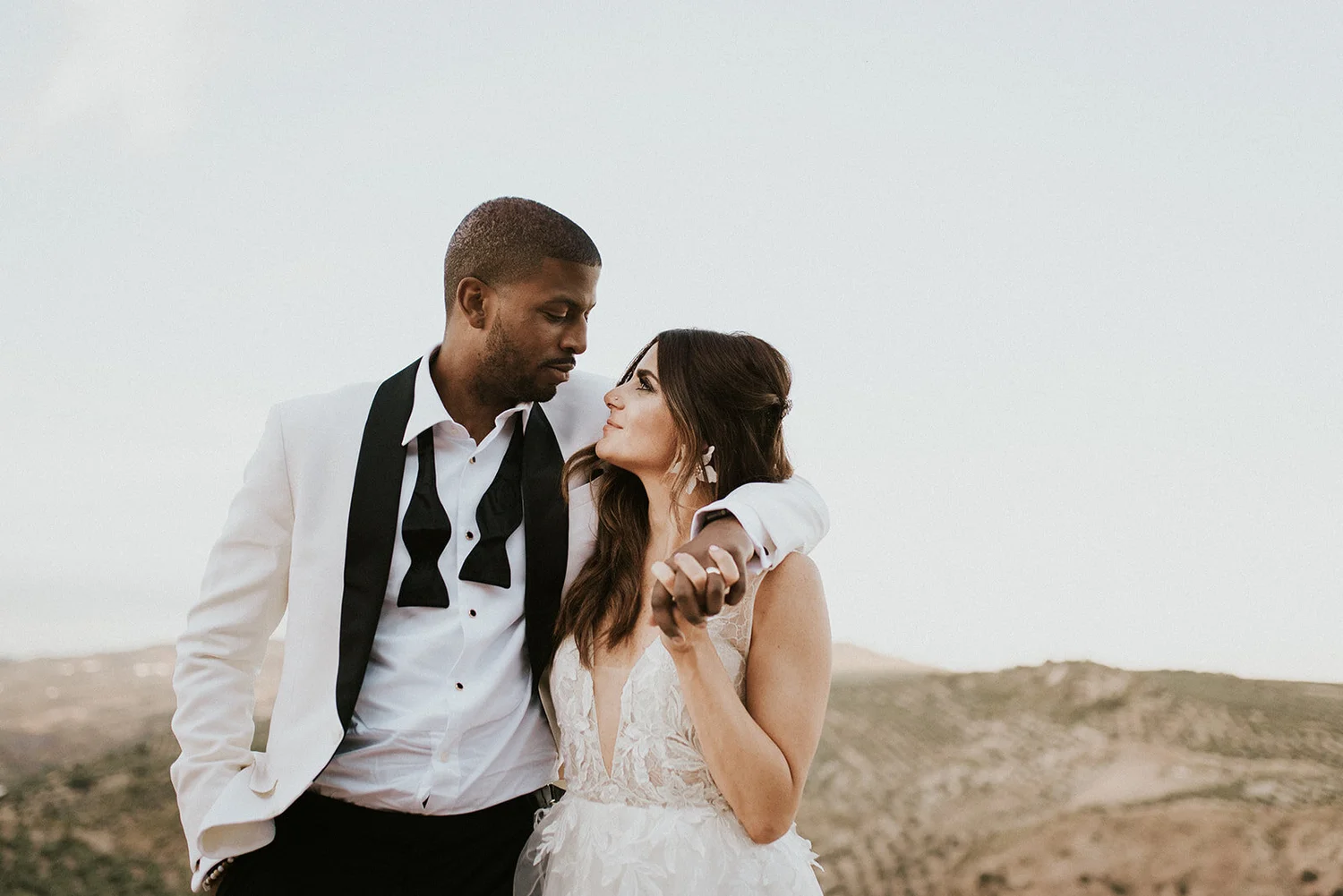 A couple dressed in wedding attire with a desert landscape in the background, gazing into each other's eyes.