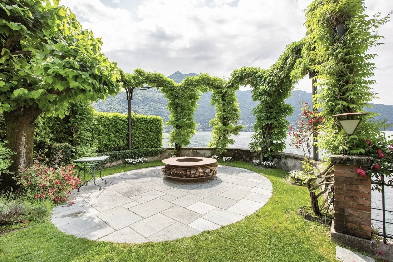 A scenic garden by a lake with green trees, bushes, and a stone fire pit on a paved circular patio, overlooking mountains under a cloudy sky.
