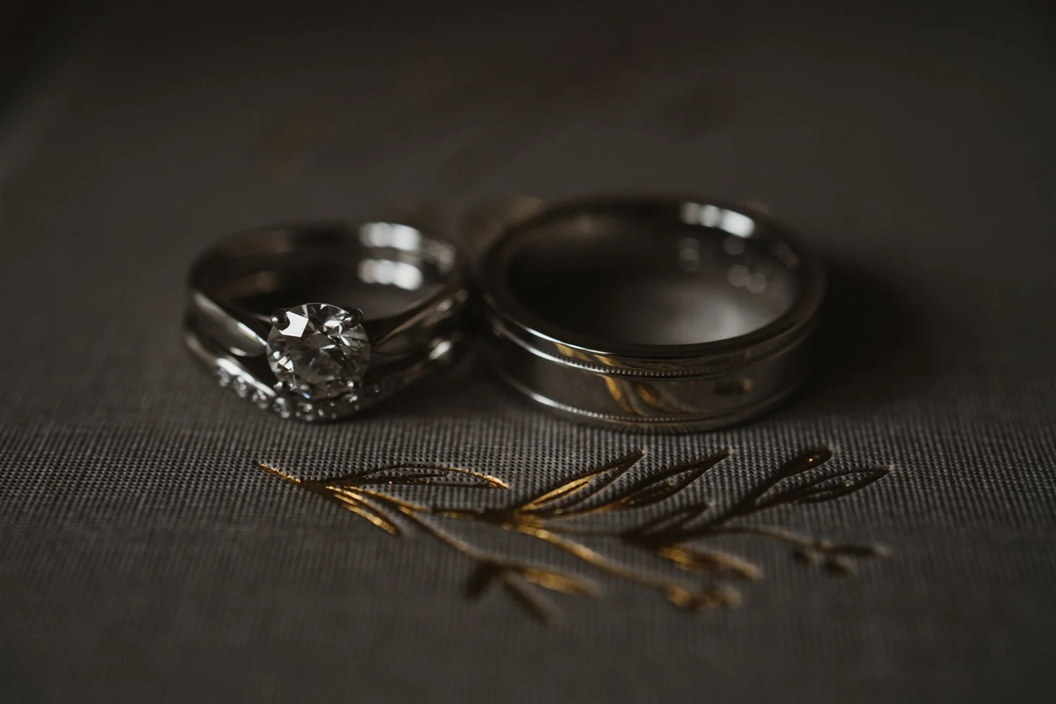 Close-up of a diamond engagement ring and wedding band, gold jewelry, and decorative golden details on a dark textured surface.
