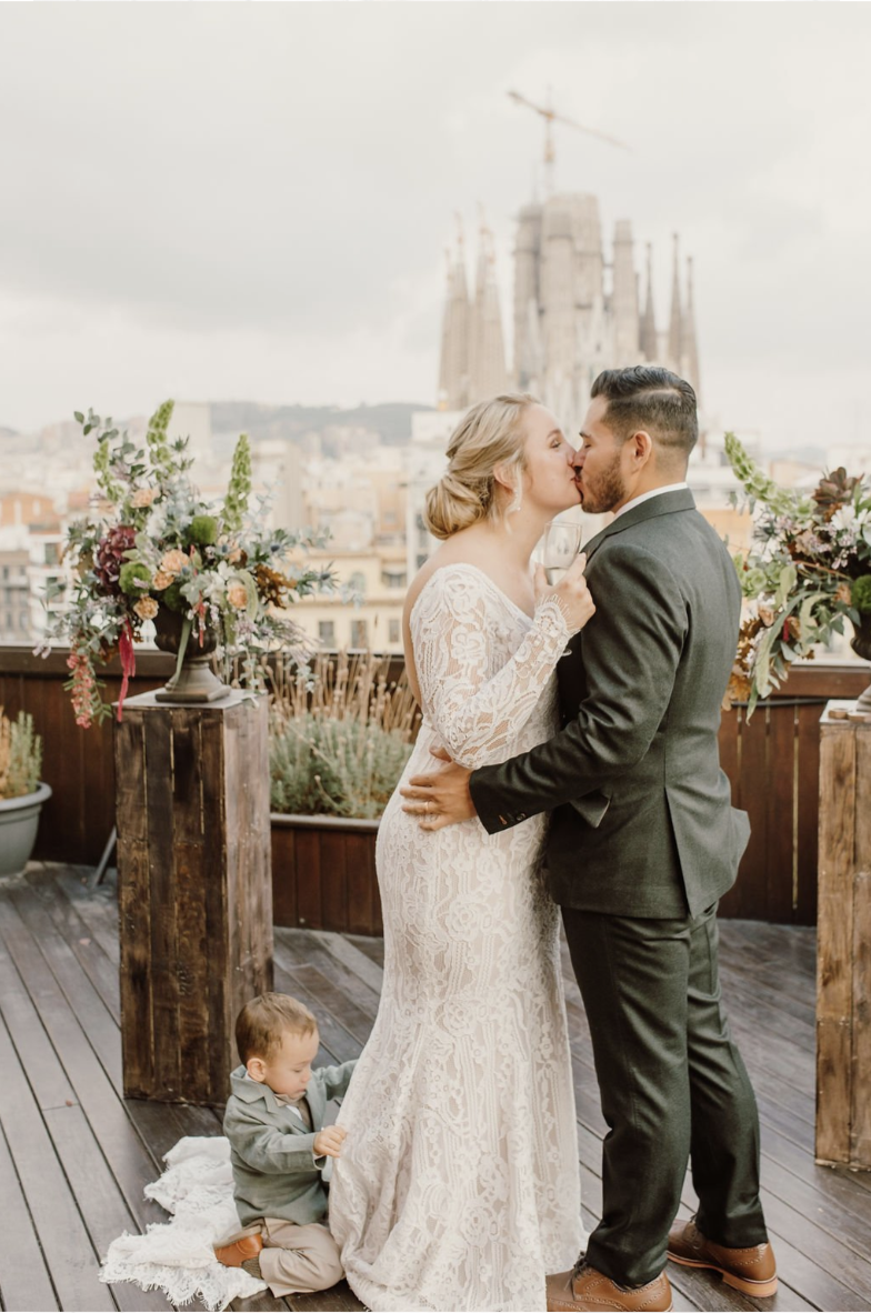 A bride and groom kiss on a rooftop balcony with city buildings and the Sagrada Familia in the background, surrounded by floral arrangements, with a small child sitting on the floor nearby.