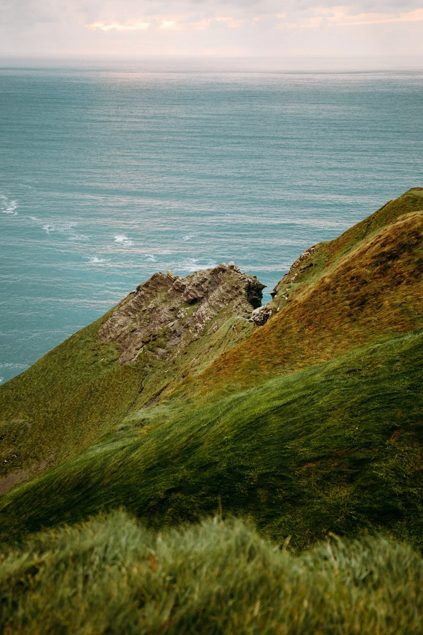 A scenic view of green rolling hills and rocky cliffs overlooking the ocean with a cloudy sky.