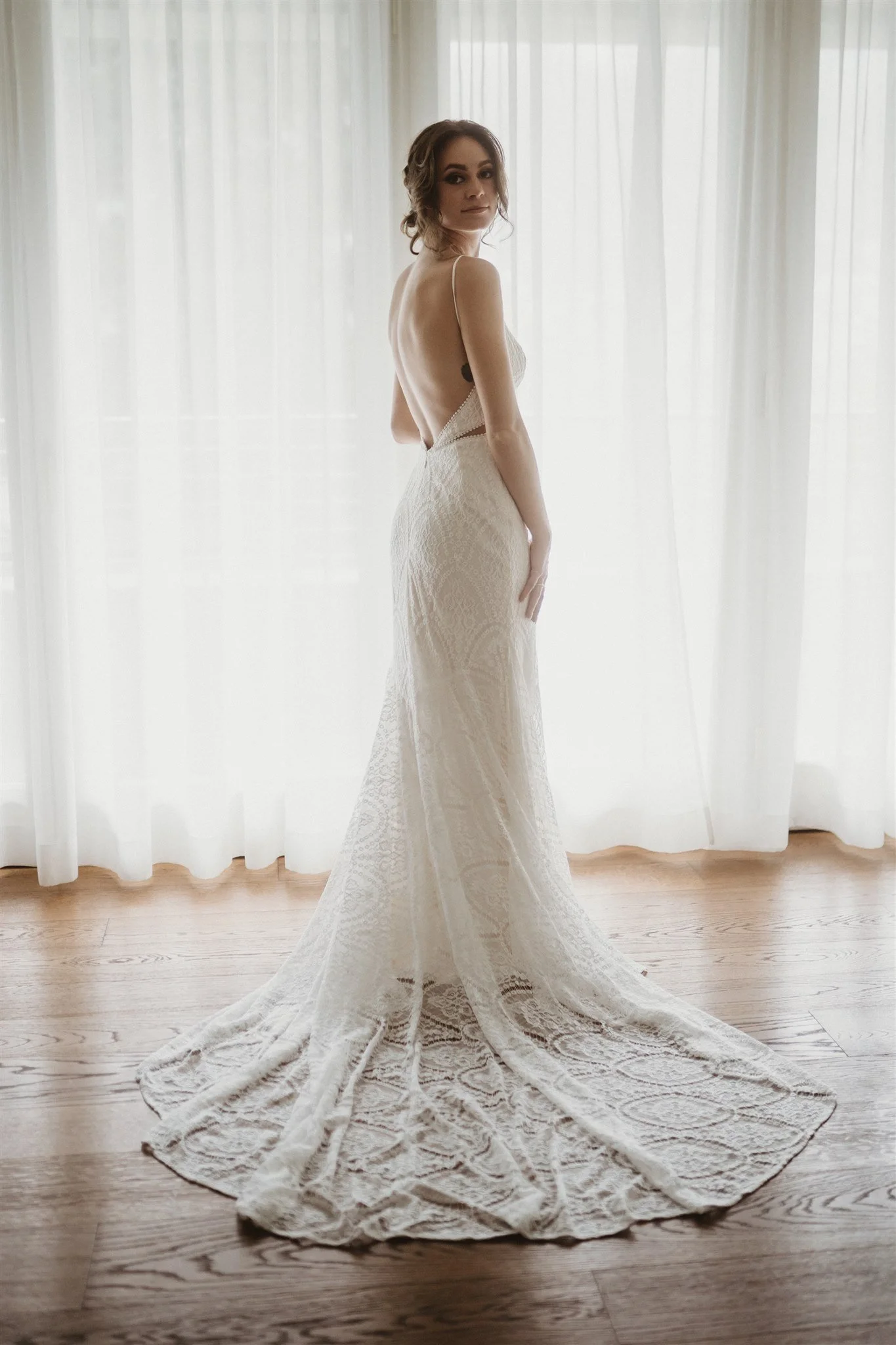 A woman in a long, white lace wedding dress standing indoors near a window with sheer white curtains, with the dress's train spread on the wooden floor.