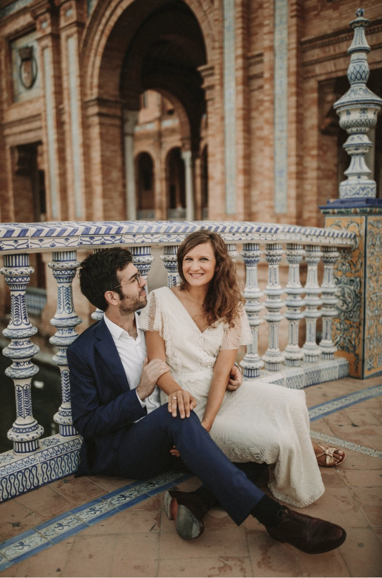 A man and woman sitting on a decorative blue and white tiled bench outside a historic brick building, smiling at each other.