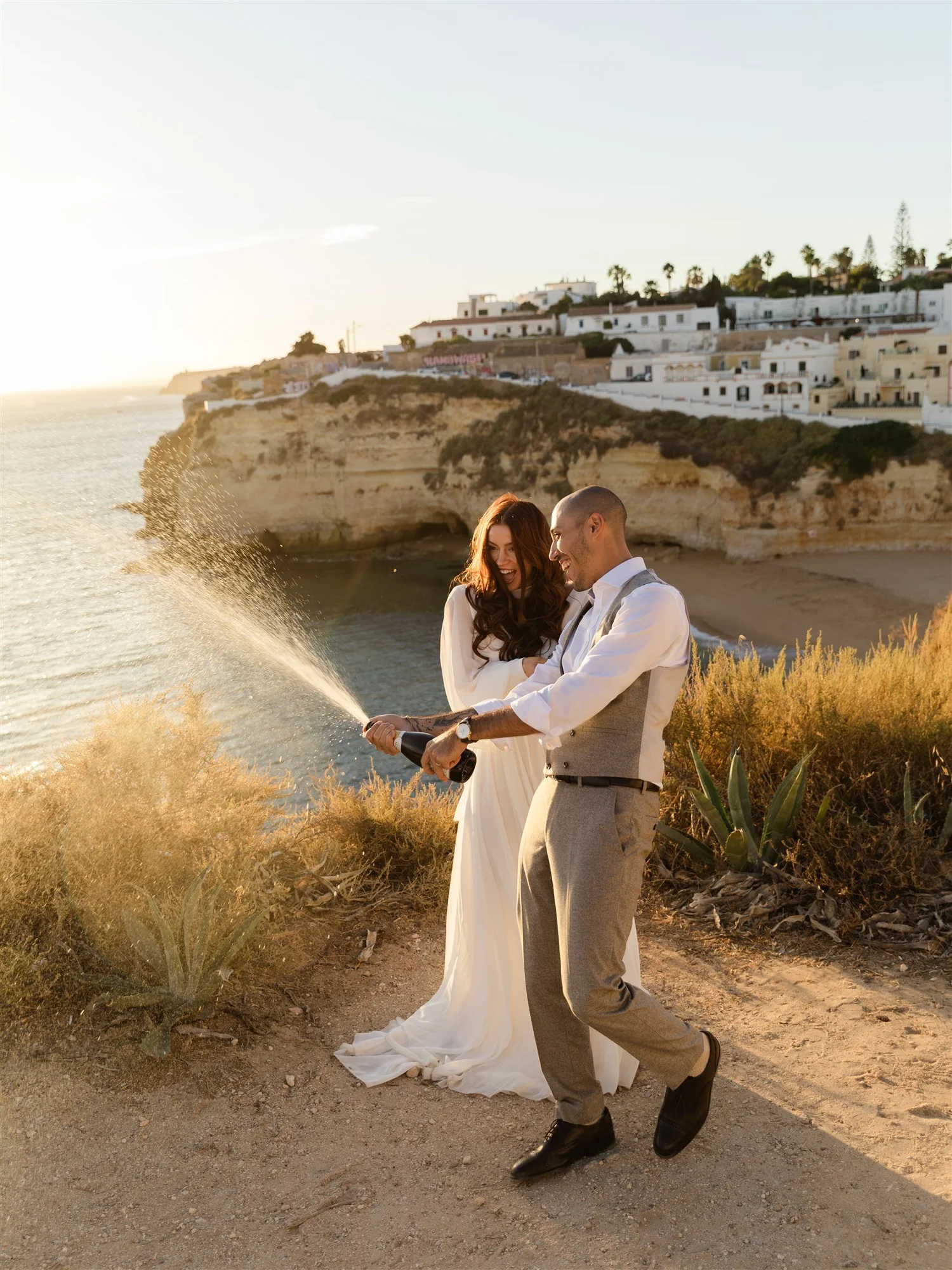 A bride and groom celebrating on a beach with cliffs and white houses in the background, spraying champagne together during sunset.