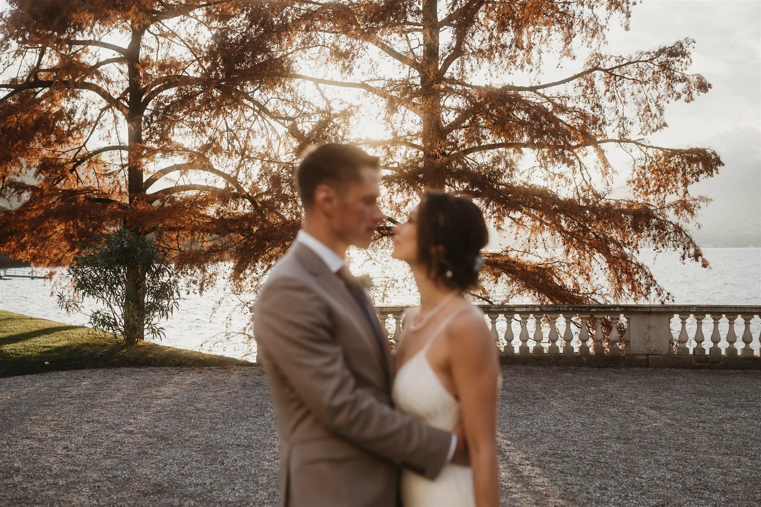 A couple dressed in wedding attire, a groom in a suit and a bride in a white dress, standing outdoors near a body of water with large trees in the background during sunset.
