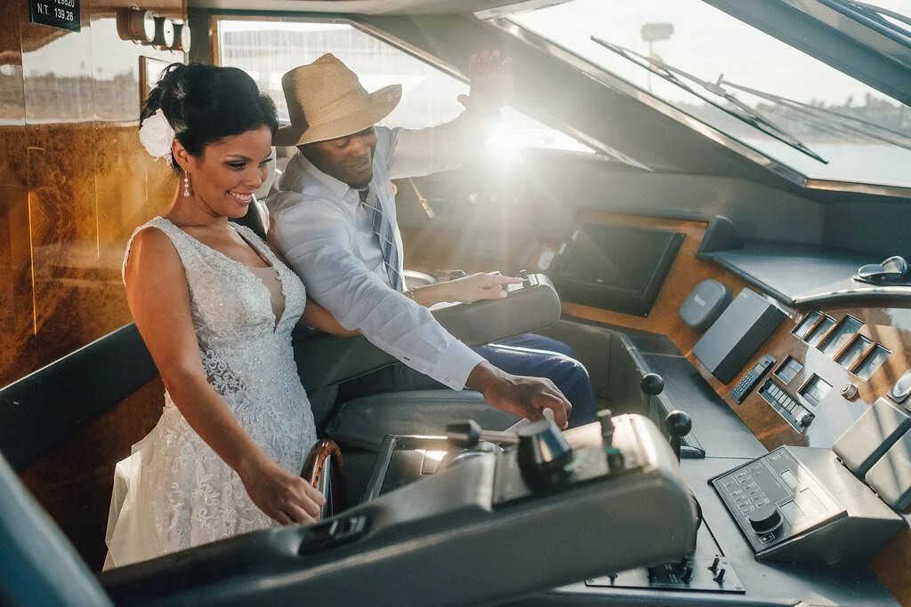 A smiling bride in a white lace wedding dress and a man in a hat sitting in the cockpit of an airplane, looking at the controls.