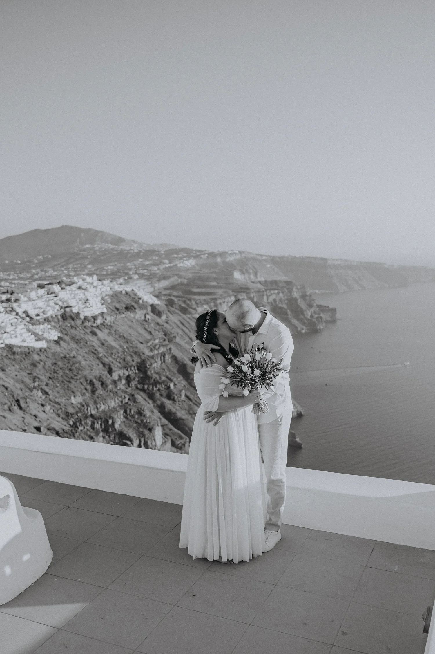 A couple, dressed in wedding attire, shares a kiss on a terrace overlooking the coast with cliffs and water in the background.