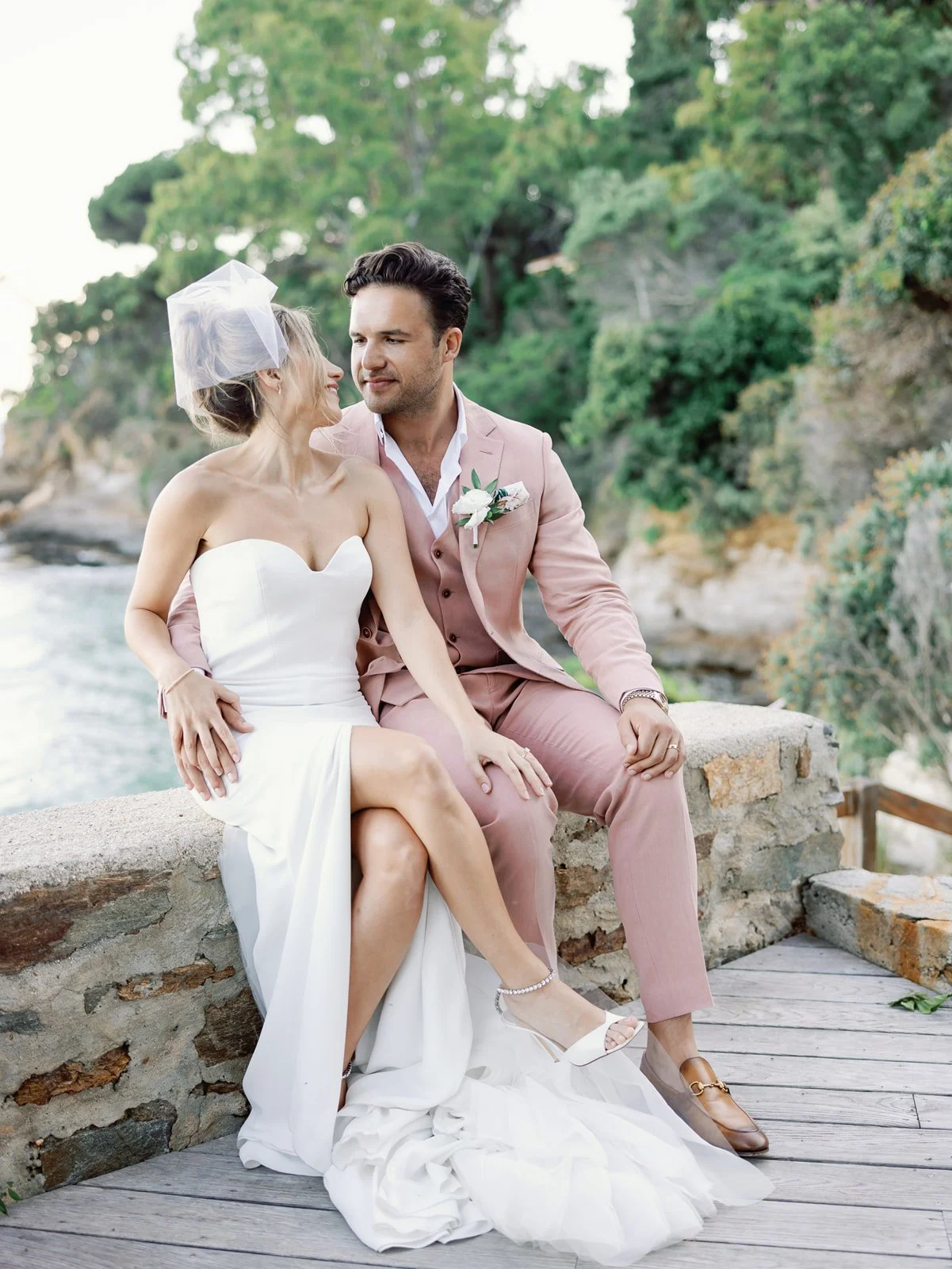 A bride and groom sitting close together on a stone wall near water and trees, during a wedding photo shoot, with the bride wearing a strapless white dress and veil, and the groom in a pink suit, holding hands and looking into each other's eyes.