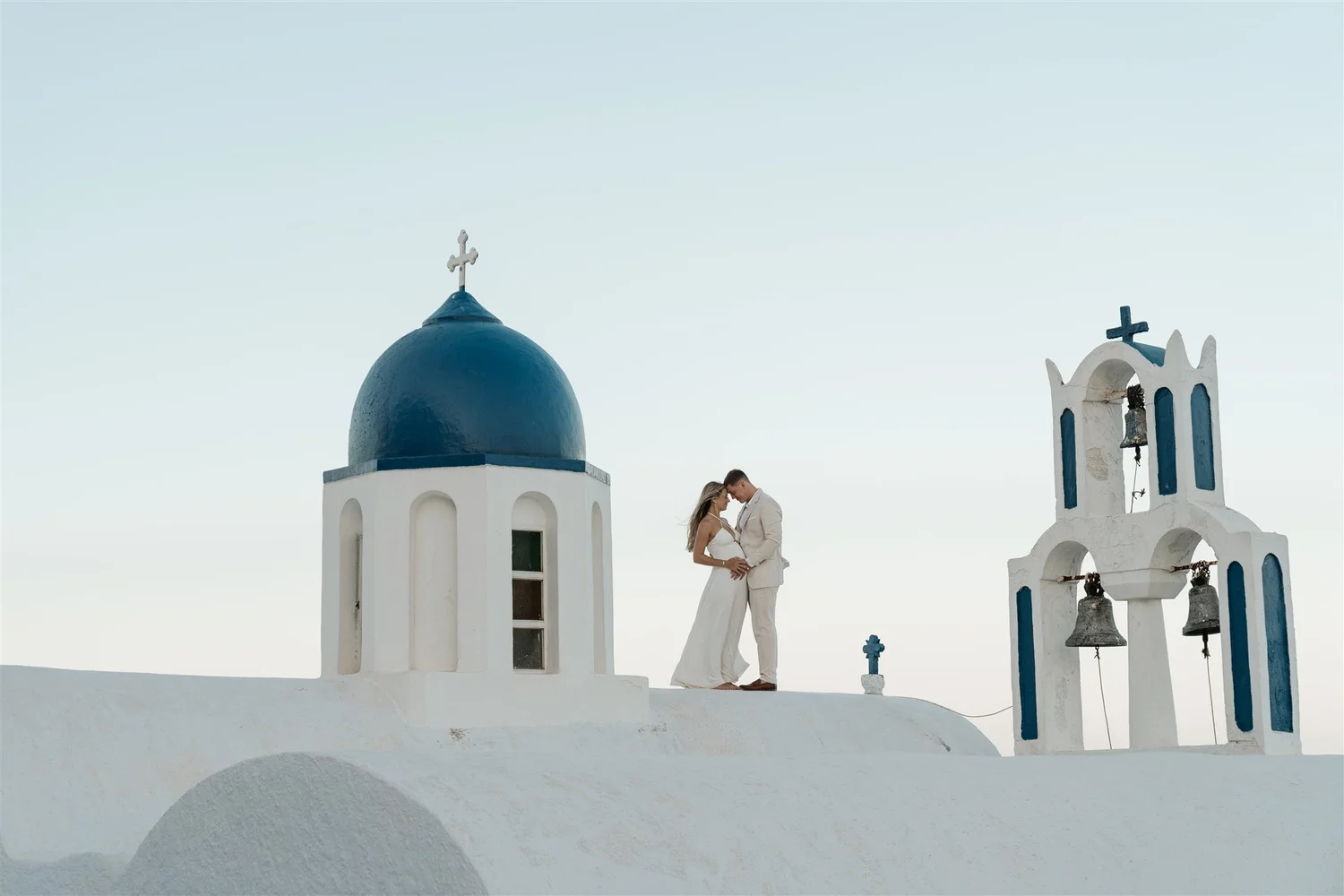 A bride and groom standing close together on a white rooftop in front of a white and blue church with bells and crosses, during what appears to be a wedding photo shoot.
