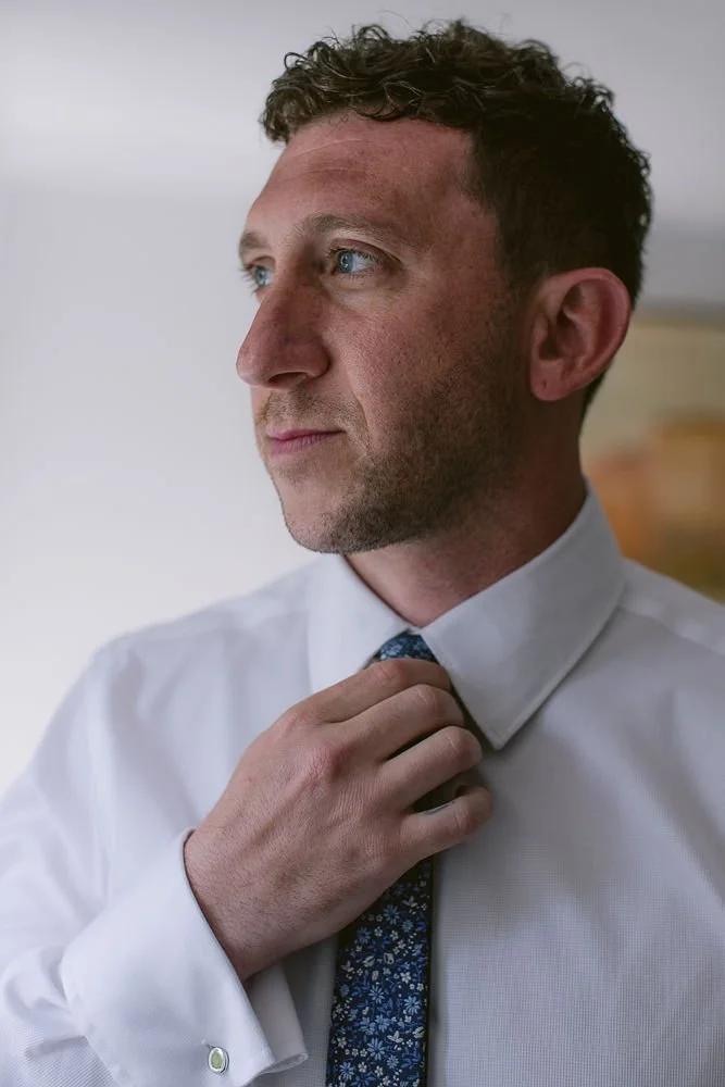 A man with curly brown hair and blue eyes adjusting his dark floral tie while wearing a white dress shirt.