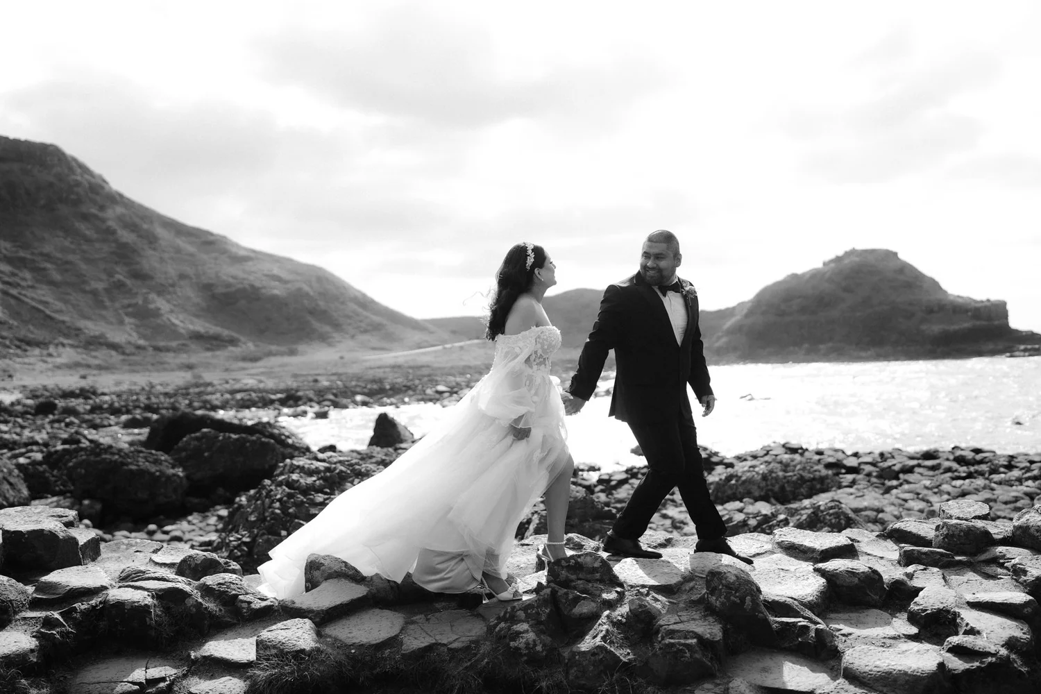 A bride and groom walk hand in hand on a rocky shoreline near the ocean, with hills in the background, captured in black and white.