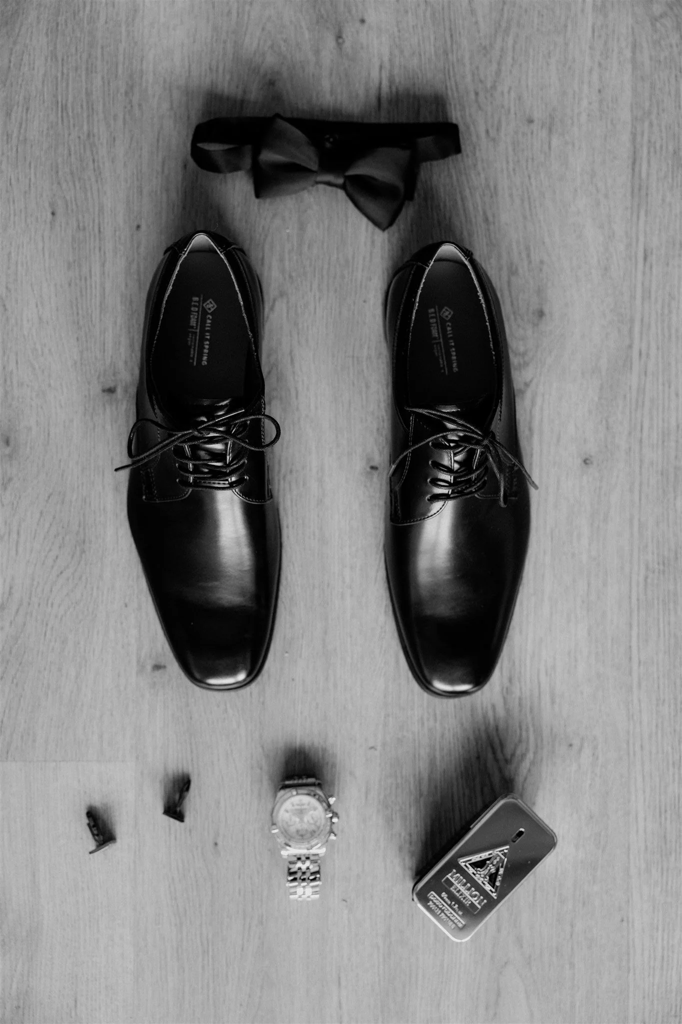Black dress shoes, a black bow tie, a wristwatch, and a pack of gum on a wooden floor, with a pair of glasses and a smartphone nearby.