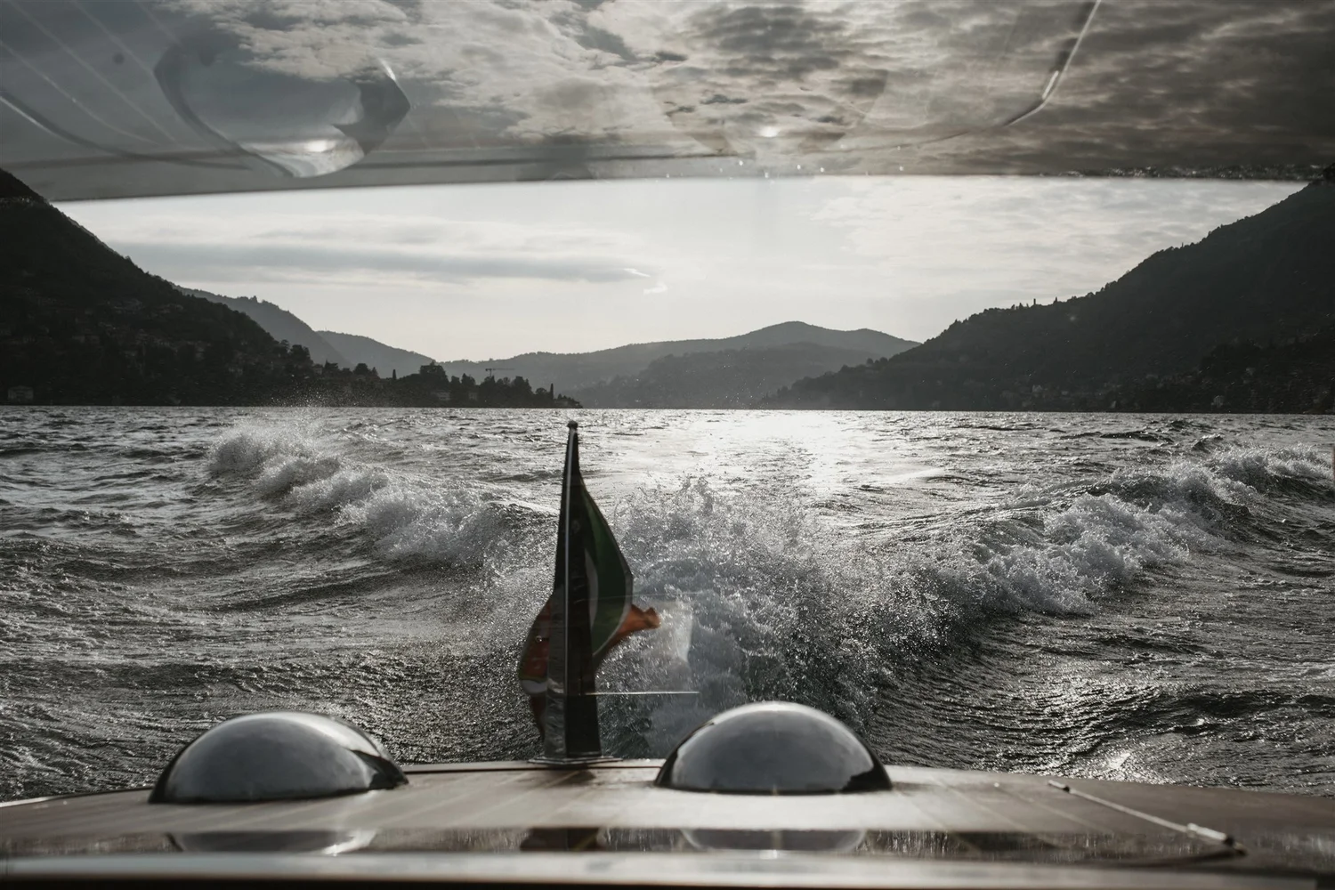 View from behind a boat on a lake with mountains in the background and a cloudy sky.