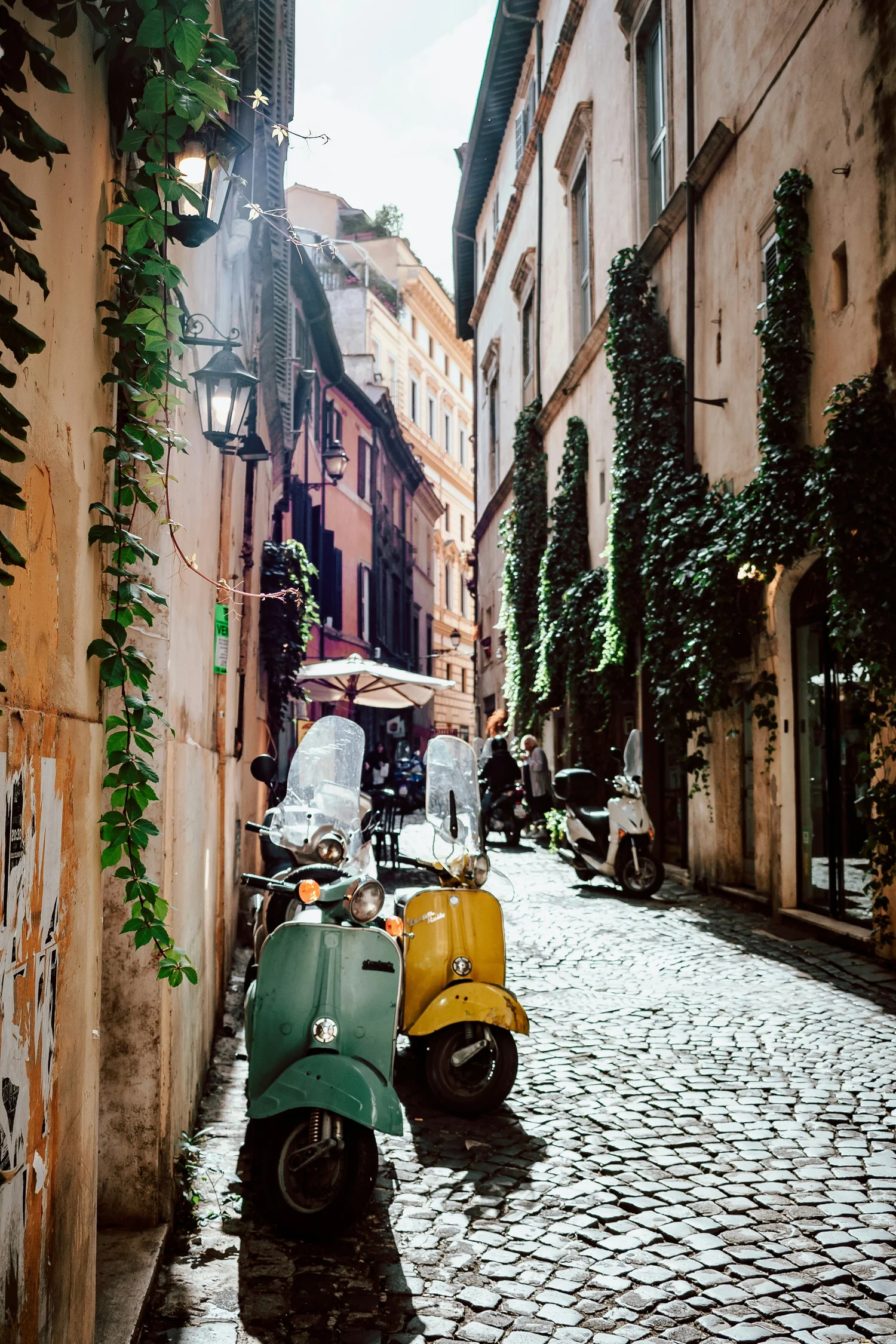 A narrow cobblestone street lined with parked scooters and motorcycles. Buildings with ivy-covered walls and vintage-style street lamps on the sides. Umbrellas and people in the distance under a bright sky.