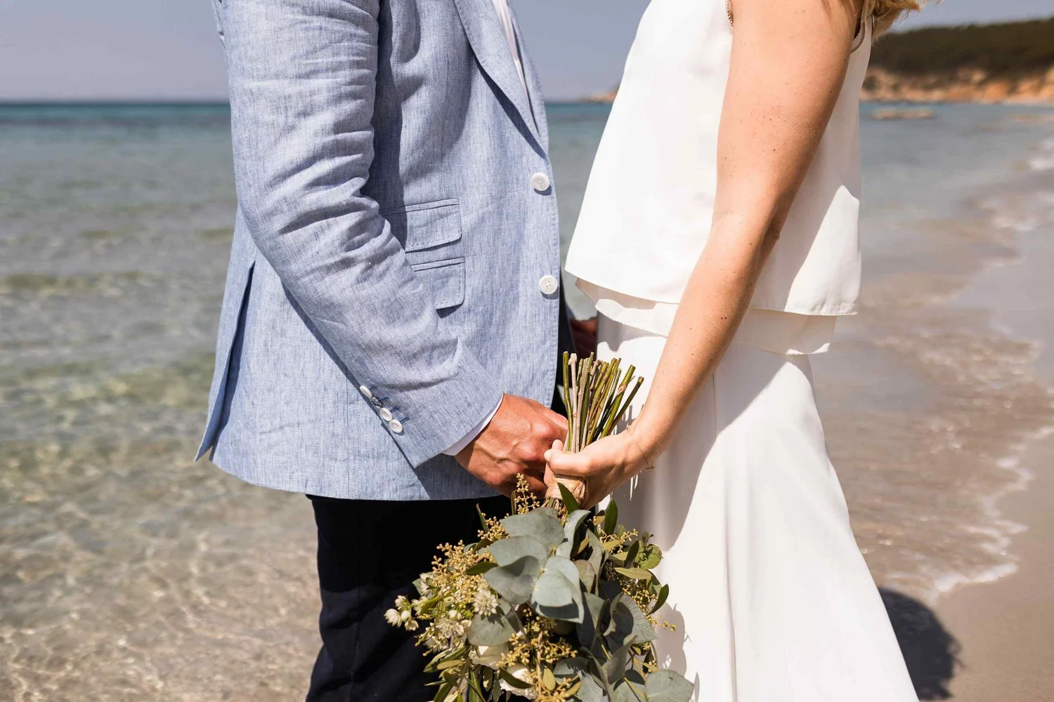 A couple holding hands on the beach, dressed in wedding attire with the man in a light blue suit jacket and the woman in a white dress, holding a bouquet of flowers.