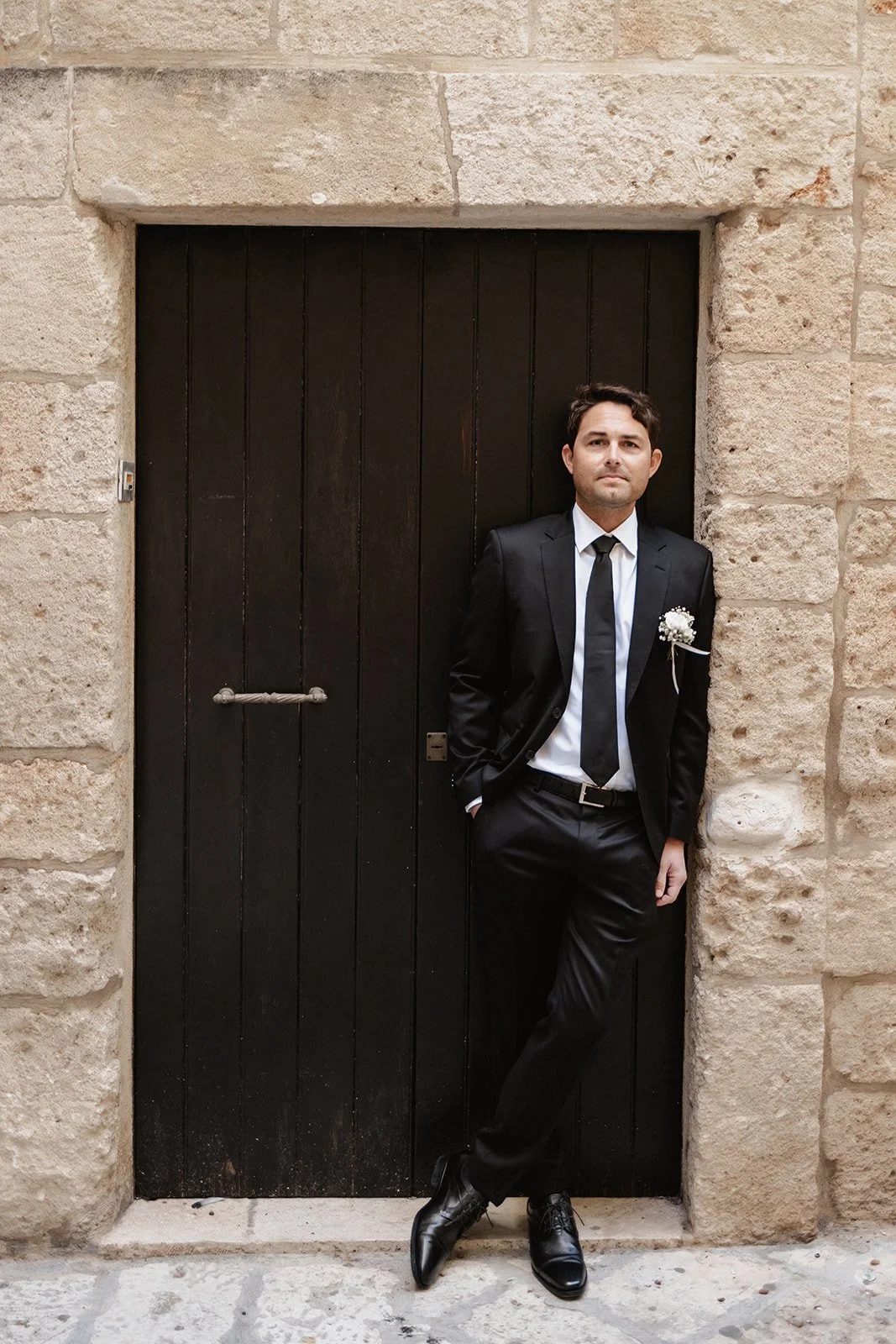 A man in a black suit and tie, with a white shirt and a boutonniere, leaning against a stone wall and door, looking at the camera.