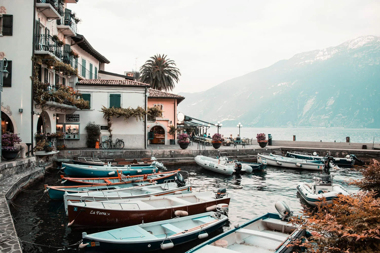 Boats docked in a marina by a paved walkway with buildings and outdoor dining tables nearby, overlooking mountains in the background during daytime.