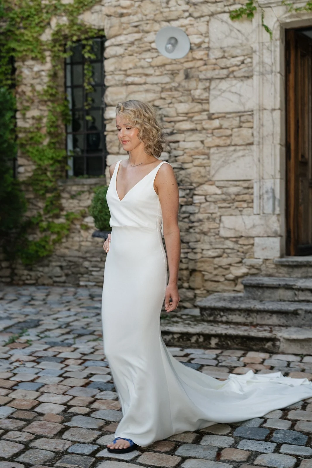 A woman in a white wedding dress walking on cobblestone pavement outdoors with a stone building and stairs in the background.