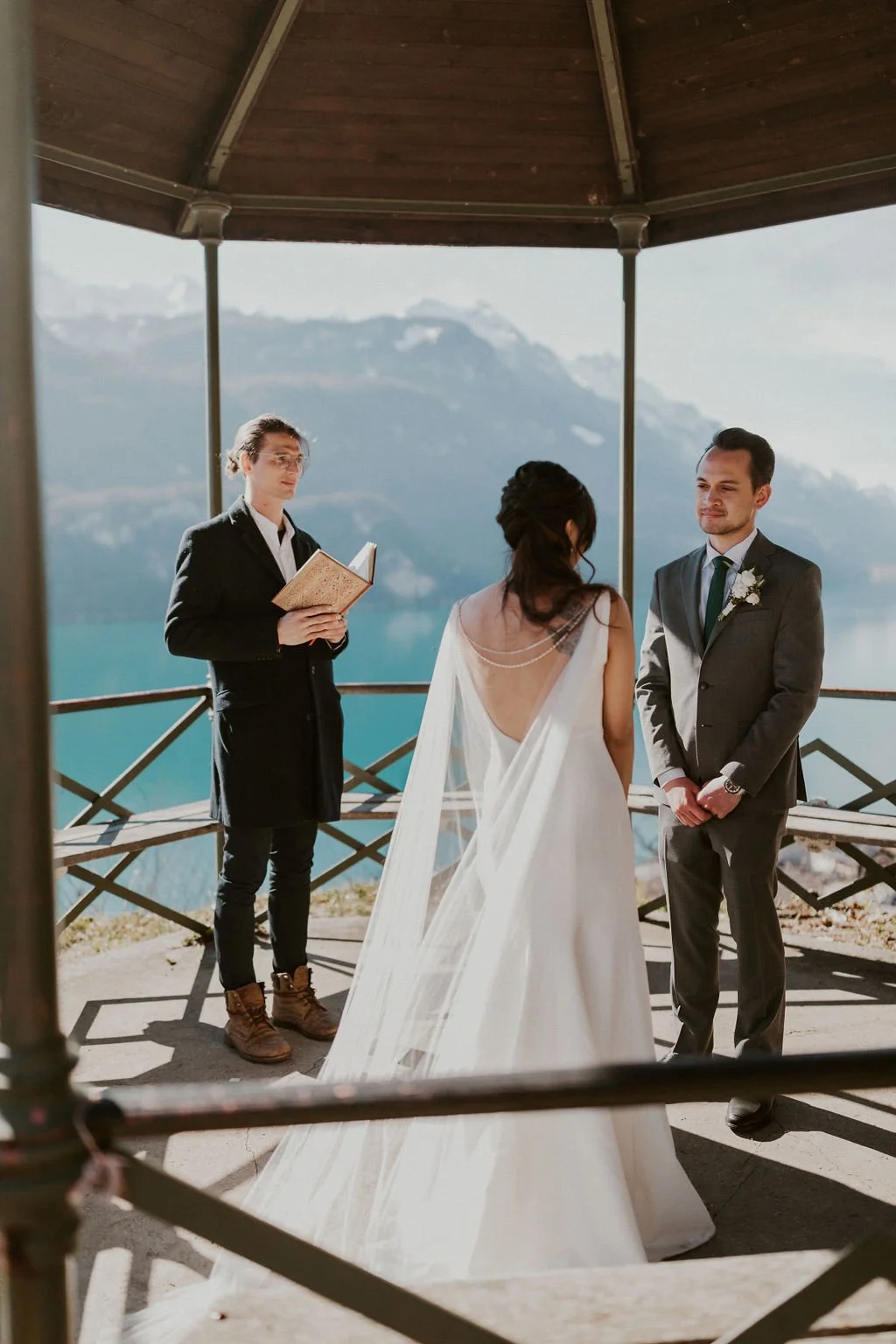 A couple is exchanging vows at an outdoor wedding ceremony with a mountain lake view backdrop, officiant holding a book in the background.