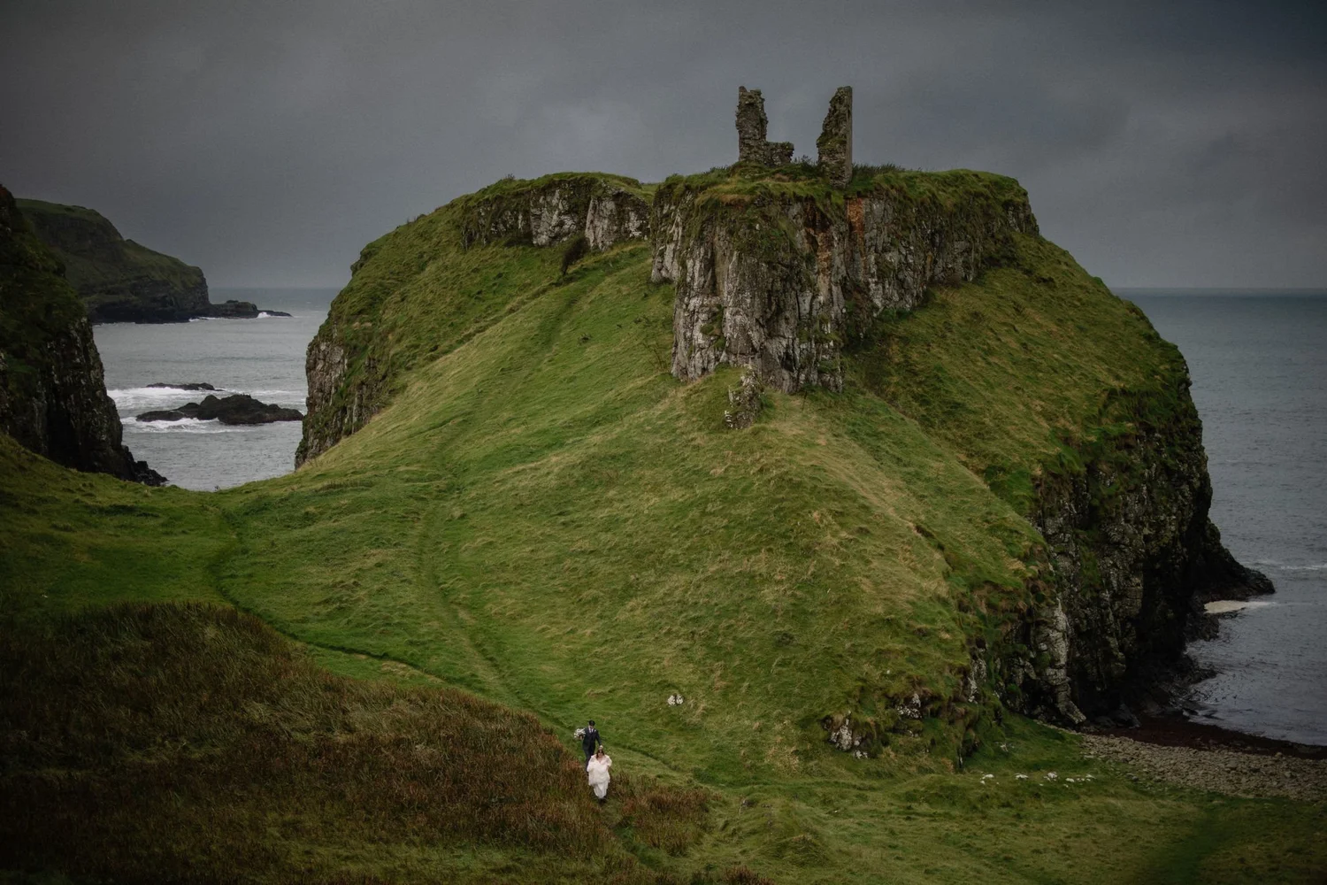 A couple dressed in wedding attire walking along a grassy hill with rugged cliffs and a castle ruin on top, overlooking the ocean on a cloudy day.