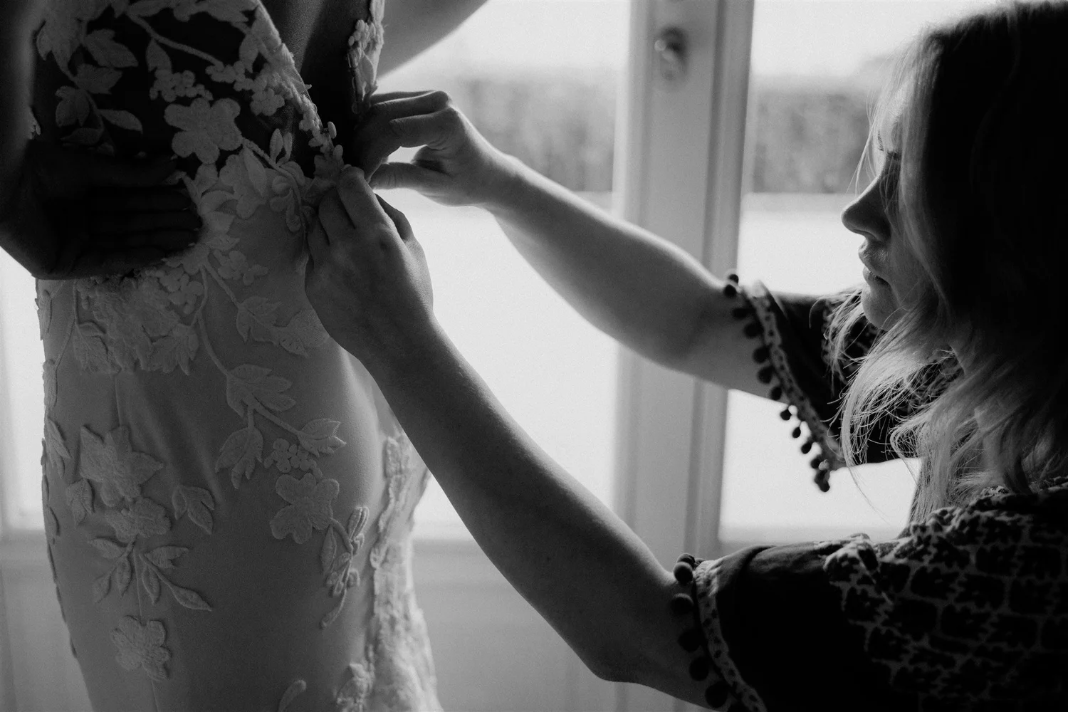 A woman adjusting the lace and floral embroidery on a wedding dress, standing by a window with natural light illuminating her profile.