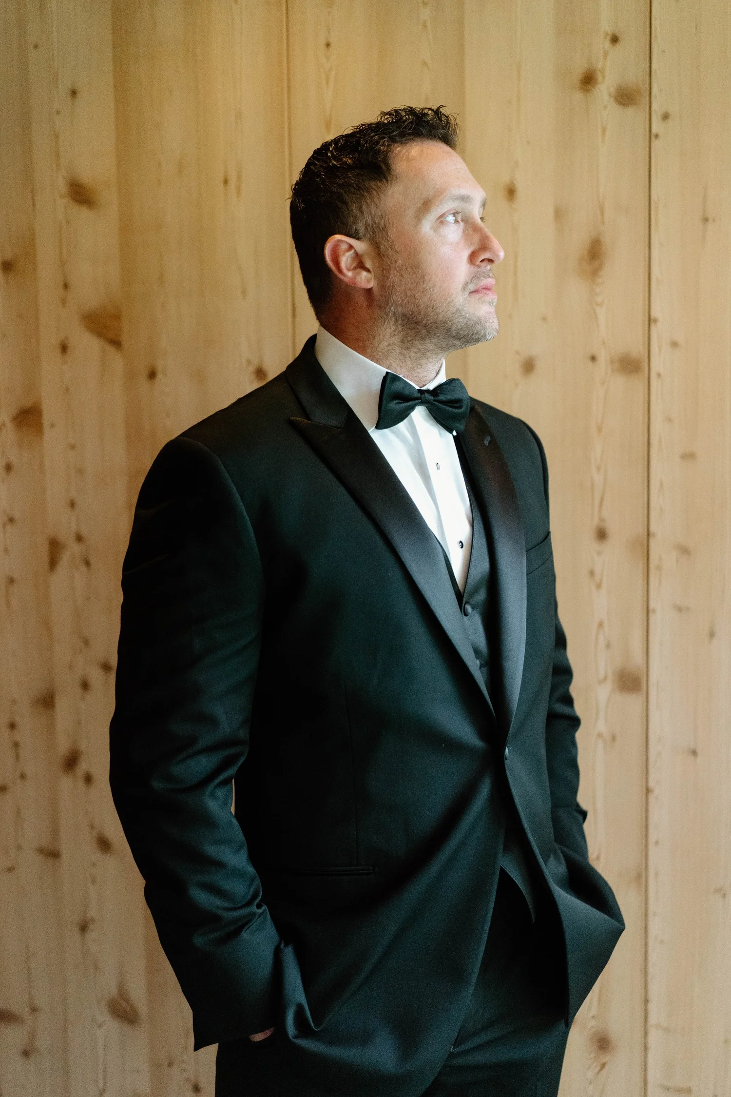 A man in a black tuxedo with a bow tie stands against a wooden wall, looking to the right.