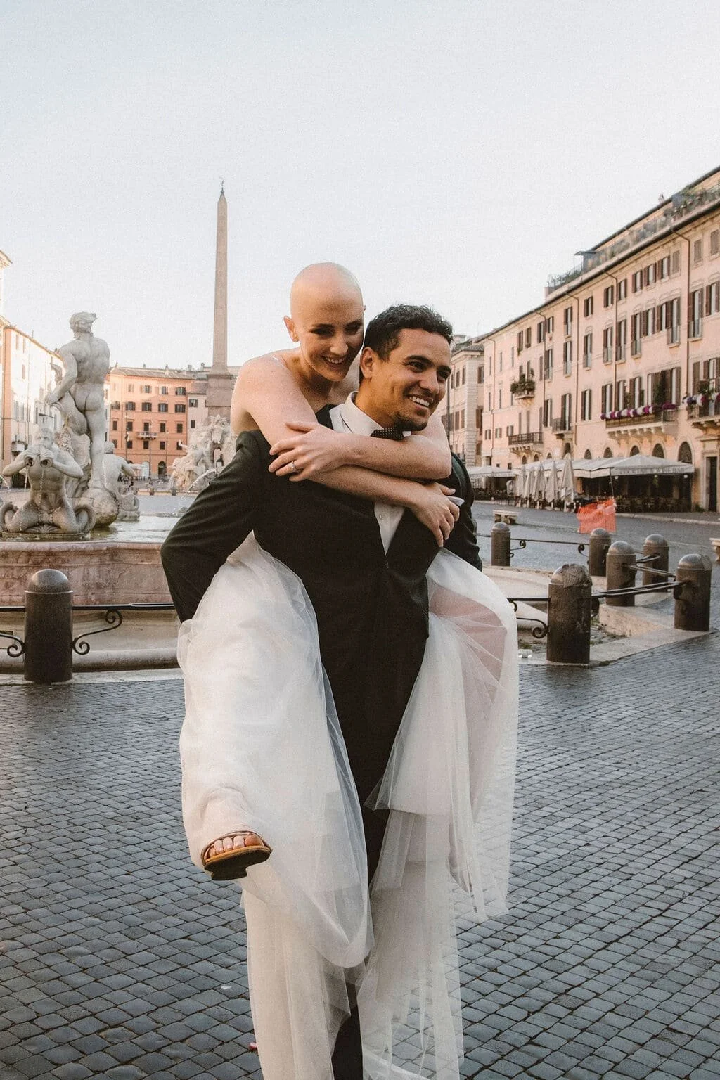 A couple, the woman with a shaved head wearing a wedding dress and the man in a tuxedo, enjoying a playful moment in a European city square, with statues and historic buildings in the background.