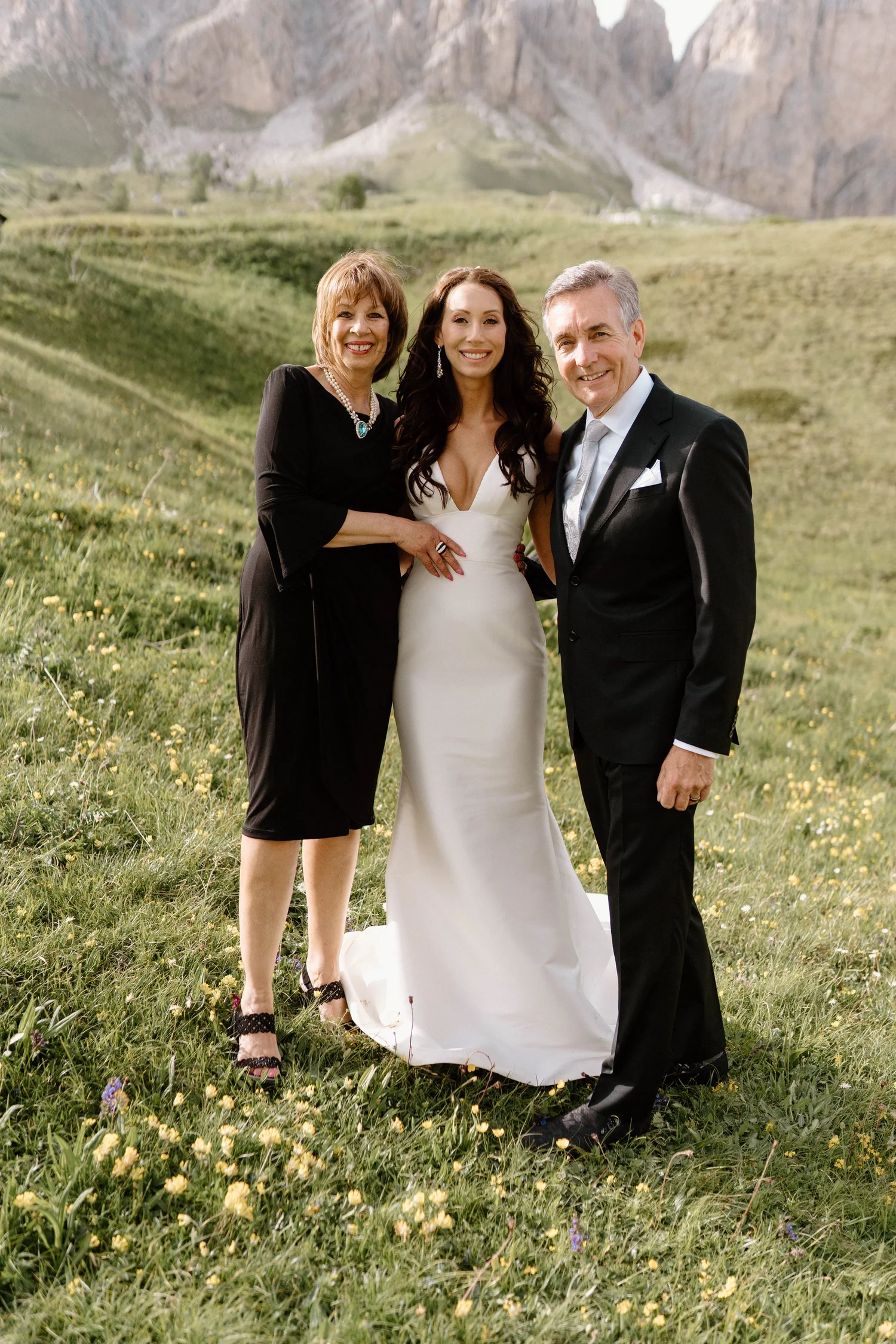 A bride in a white dress standing outdoors with a woman in a black dress and a man in a black suit, all smiling, with a mountain landscape in the background.