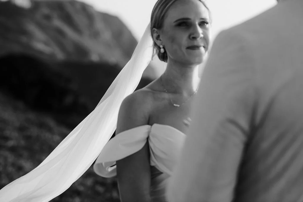 A woman in a wedding dress and veil looking at a person in casual clothing outdoors in a mountainous area.