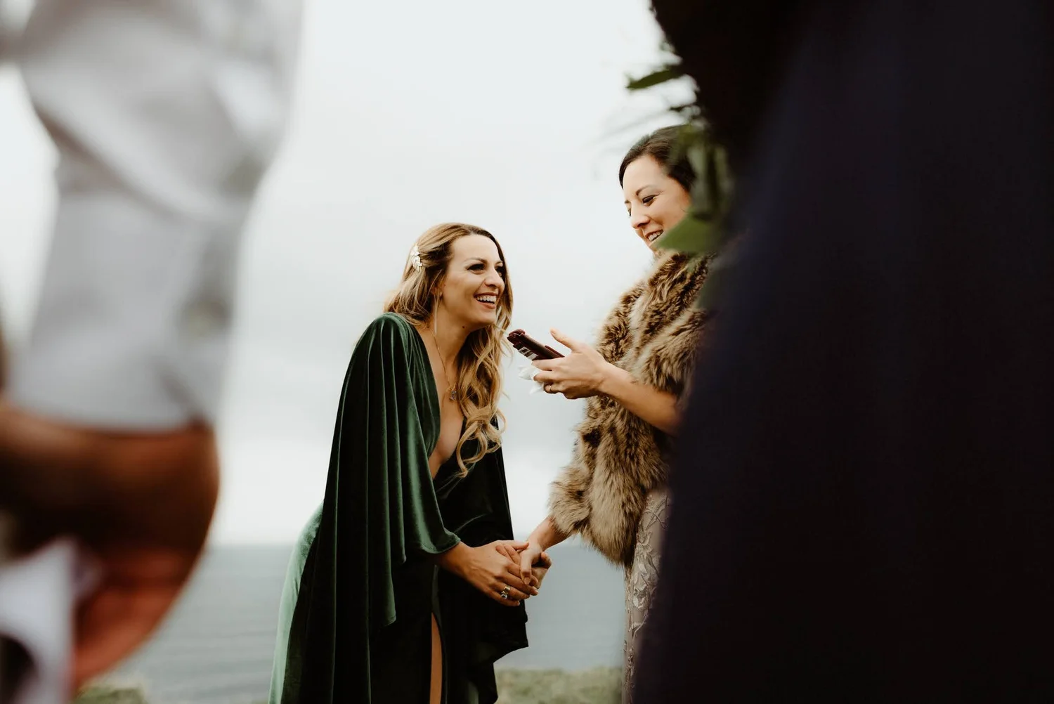 Two women dressed in formal attire are holding hands and smiling at each other during a wedding ceremony, with one woman wearing a green dress and the other wearing a jacket with fur lining.