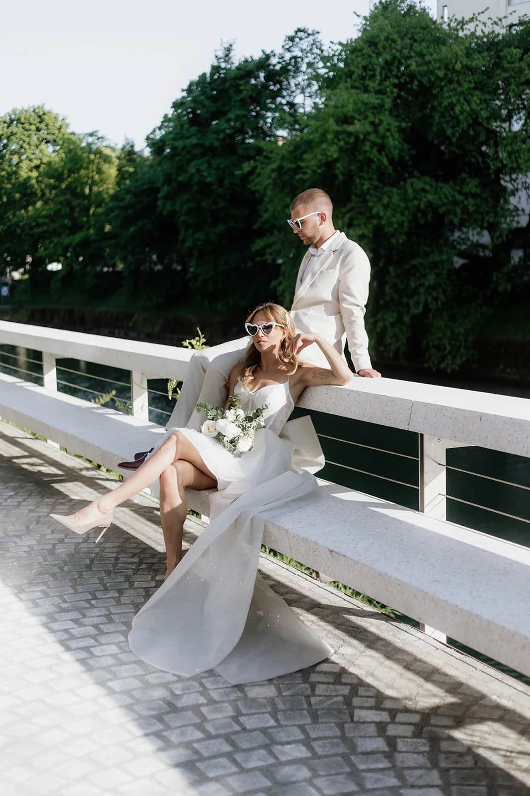 A stylish bride and groom in white outfits posing on a white bridge outdoors, with the bride sitting on the bench with her legs crossed and holding a bouquet, and the groom standing behind her, both wearing white sunglasses, surrounded by greenery.