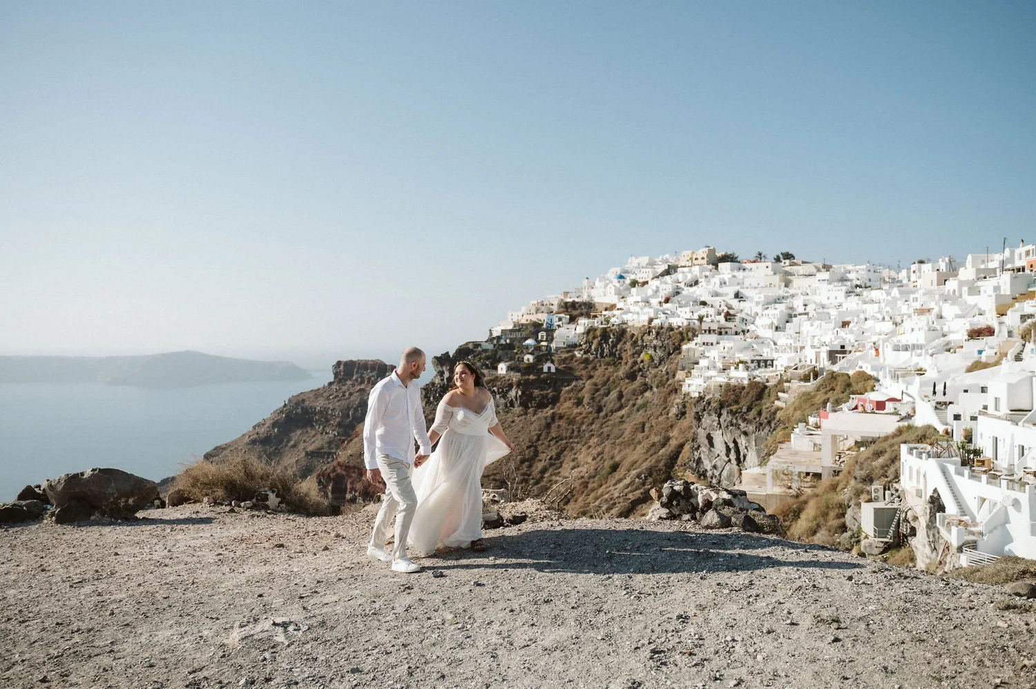 A couple in white clothes walking along a rocky path on a cliffside with white buildings on the hillside in the background.