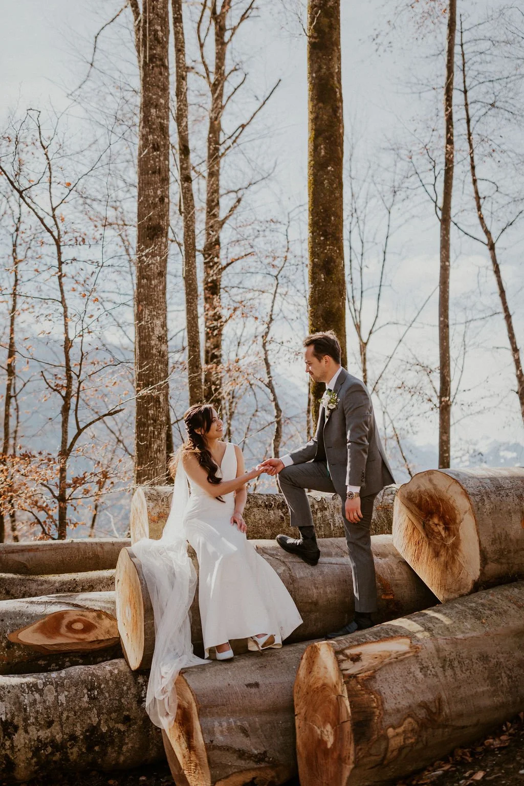 A bride and groom seated on cut logs in a forest, holding hands amidst tall, leafless trees on a bright day.