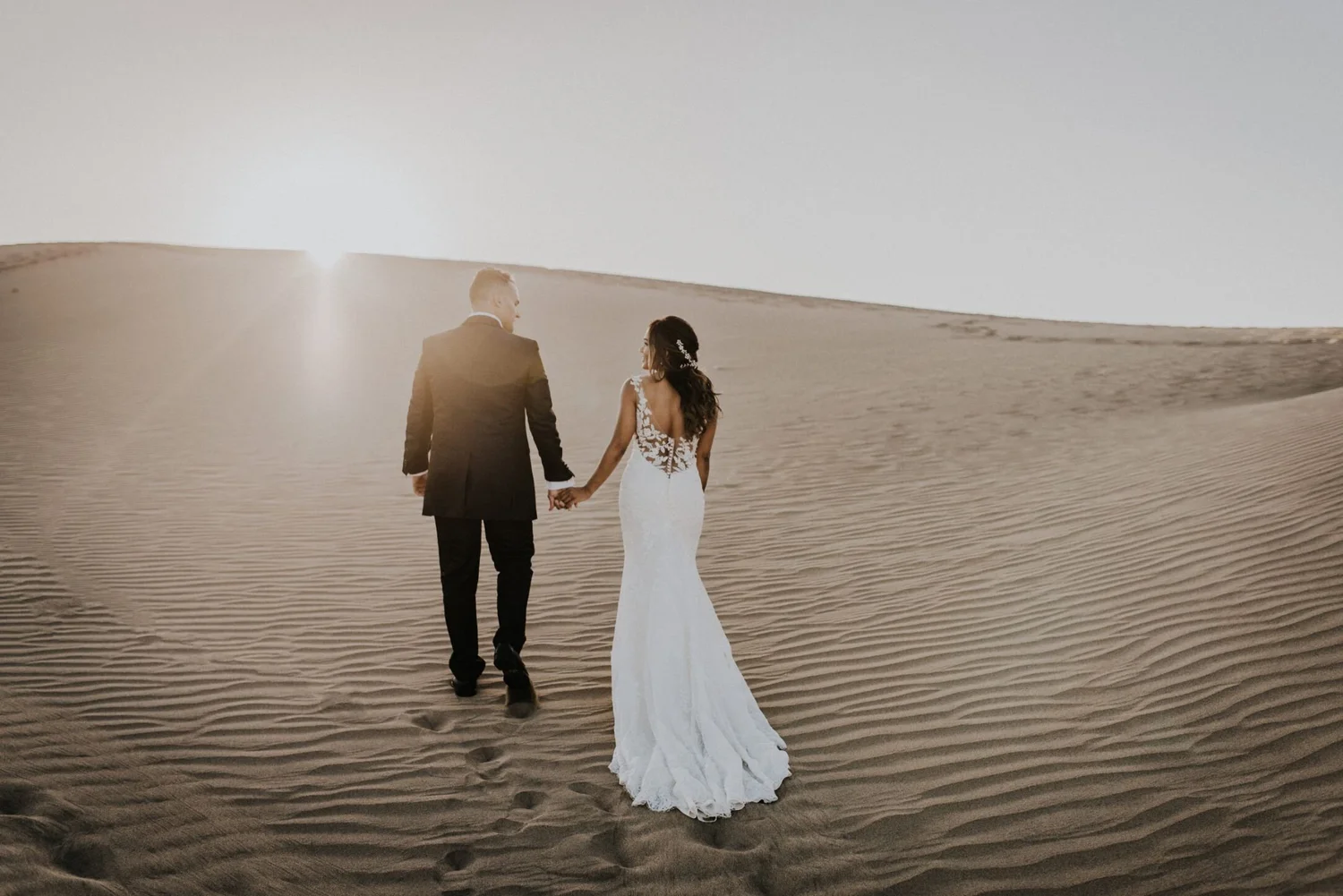 A bride and groom holding hands and walking in the desert during sunset, with the bride wearing a white wedding dress and the groom in a dark suit.