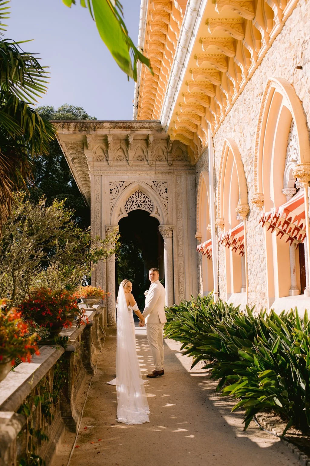 Bride and groom holding hands and facing each other outdoors in front of an ornate building with arches, greenery, and flowers.