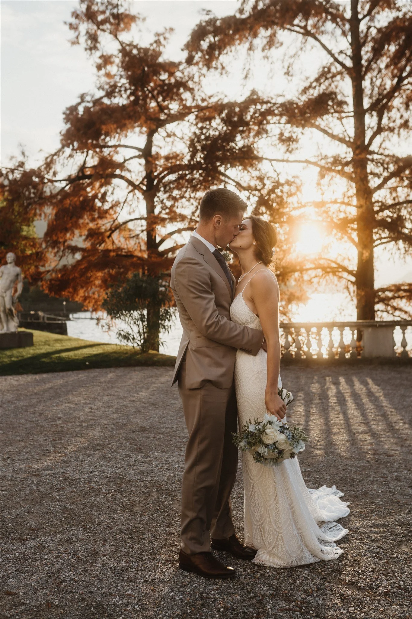 A bride and groom share a kiss outdoors during sunset, with large orange leaves on trees in the background and a body of water. The bride holds a bouquet of white flowers. The man wears a tan suit and the woman a white lace wedding dress.