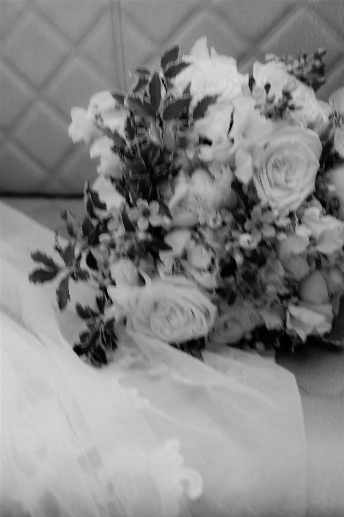 A black-and-white photograph of a bouquet of roses with variegated leaves, resting on a table or surface covered with a sheet of paper or fabric.