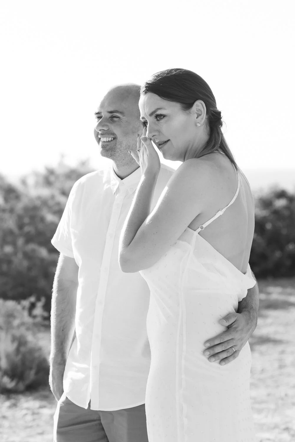 A man and woman standing outdoors, with the woman whispering into the man's ear, both smiling. Black-and-white photograph.