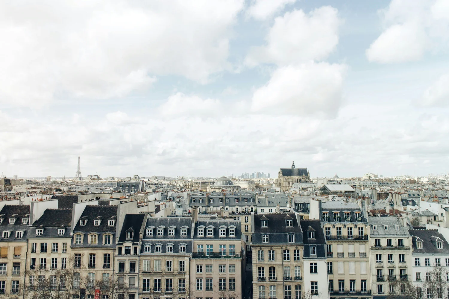 City rooftops with historic buildings and the Eiffel Tower in the distance under a cloudy sky