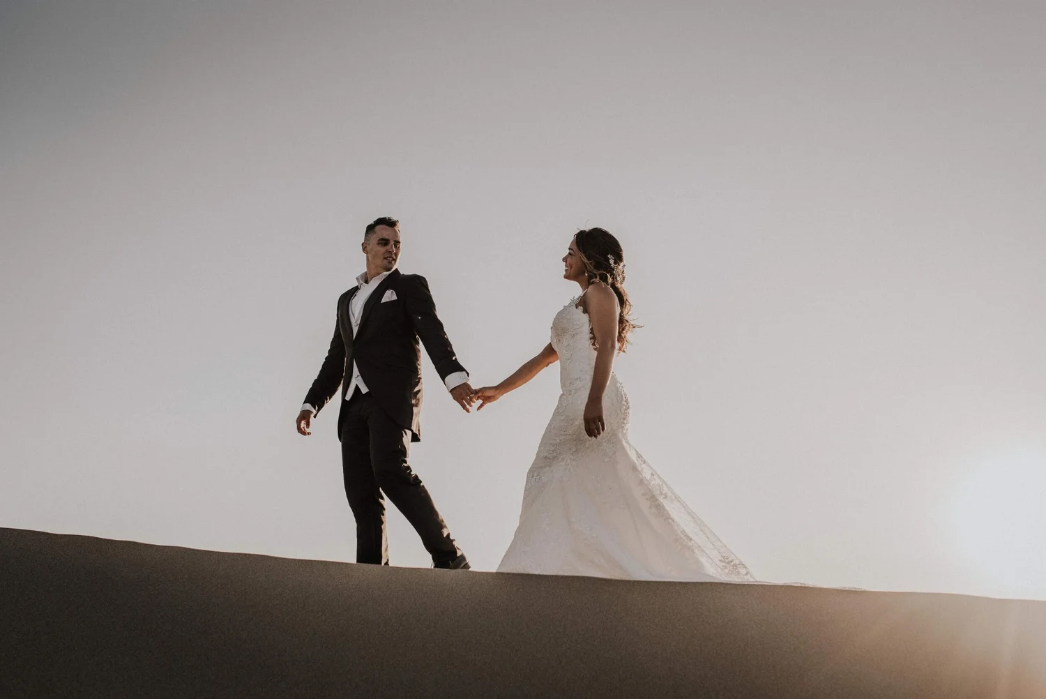 A bride and groom holding hands, standing on a curve of a sand dune against a clear sky during sunset or sunrise.