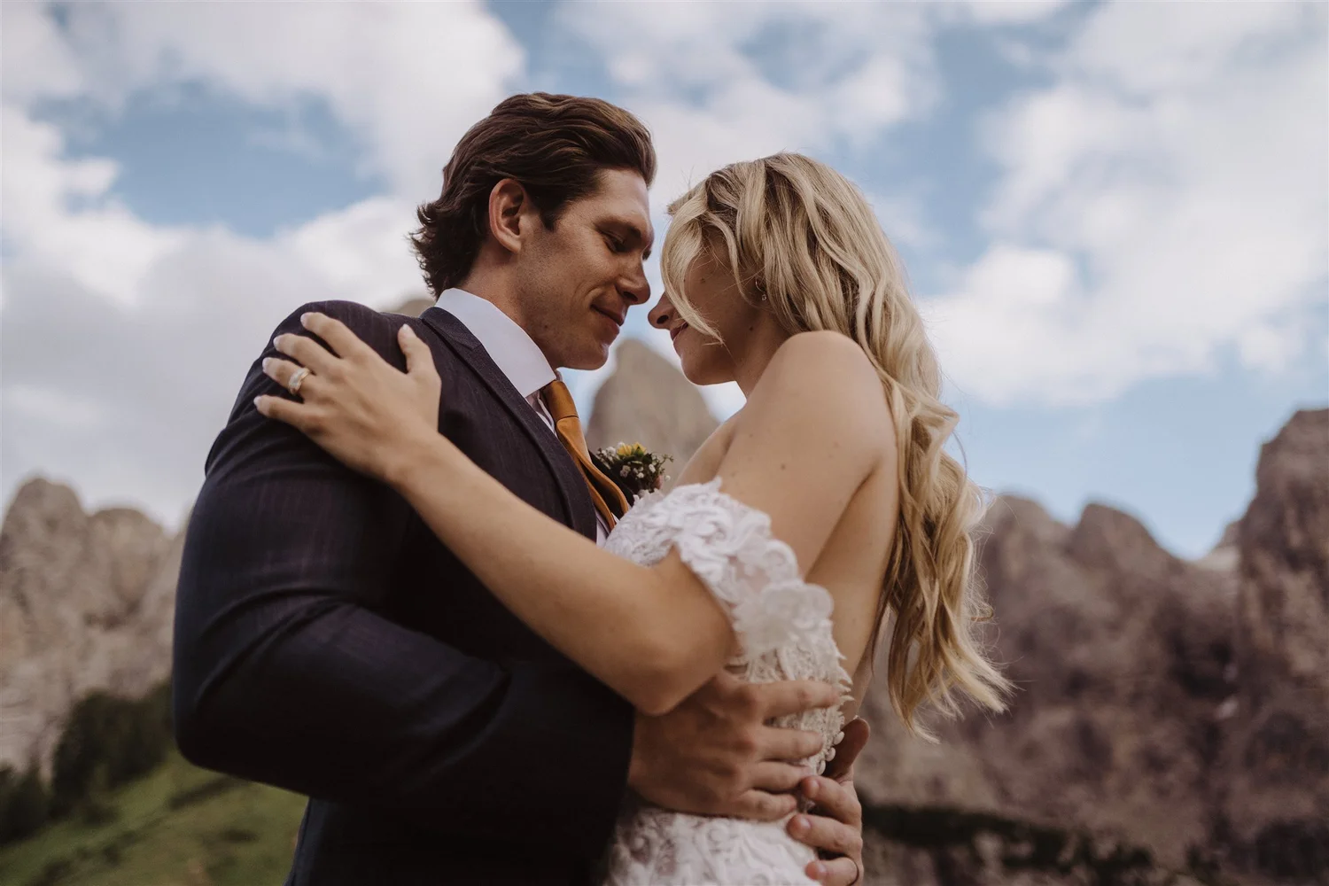 A bride and groom embrace outdoors on a cloudy day, with rocky terrain in the background.