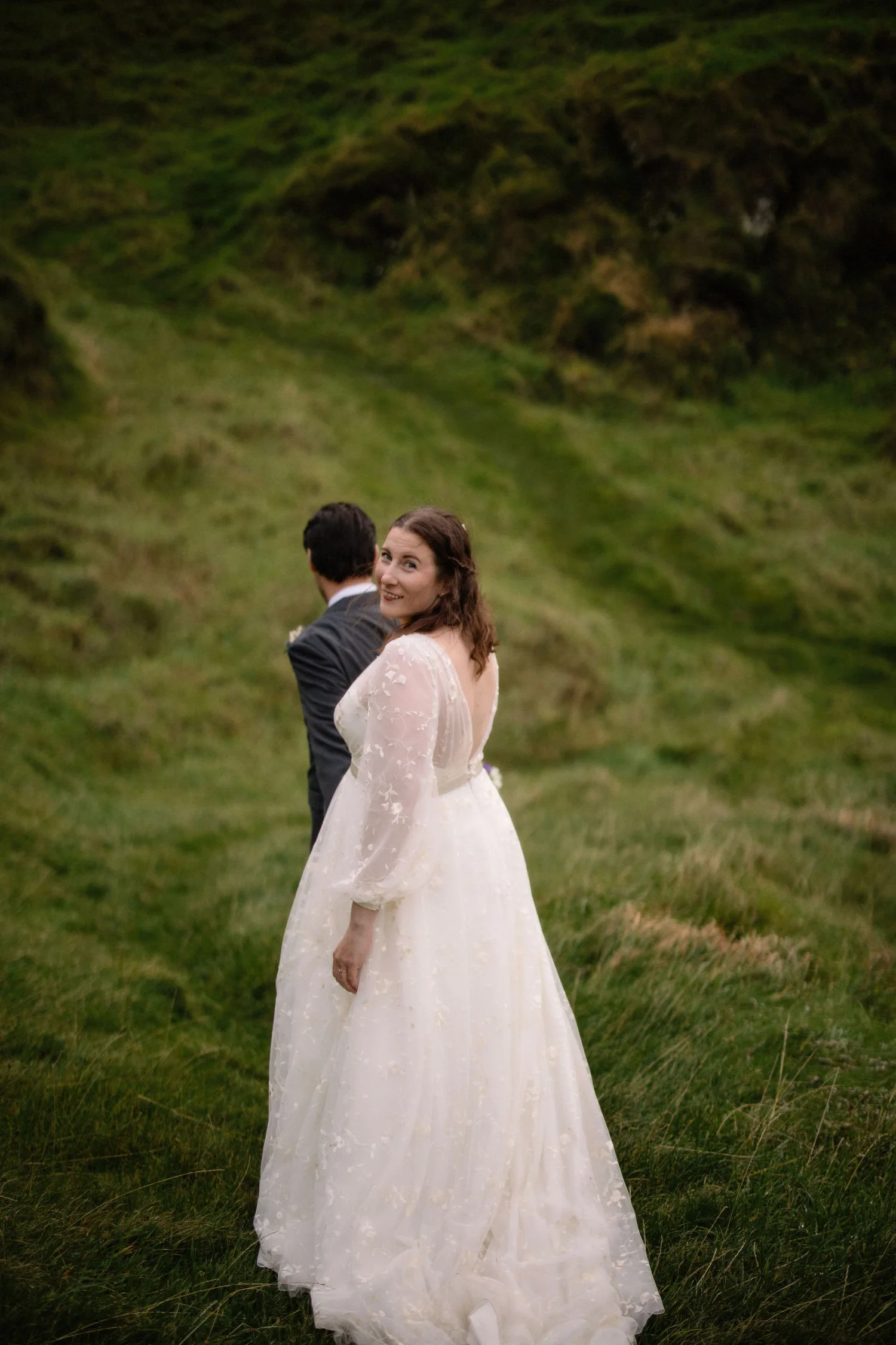 A woman in a white wedding dress standing in a grassy outdoor field with rolling green hills. She is smiling and looking back at the camera, while a man in a suit stands behind her with his back to the camera.
